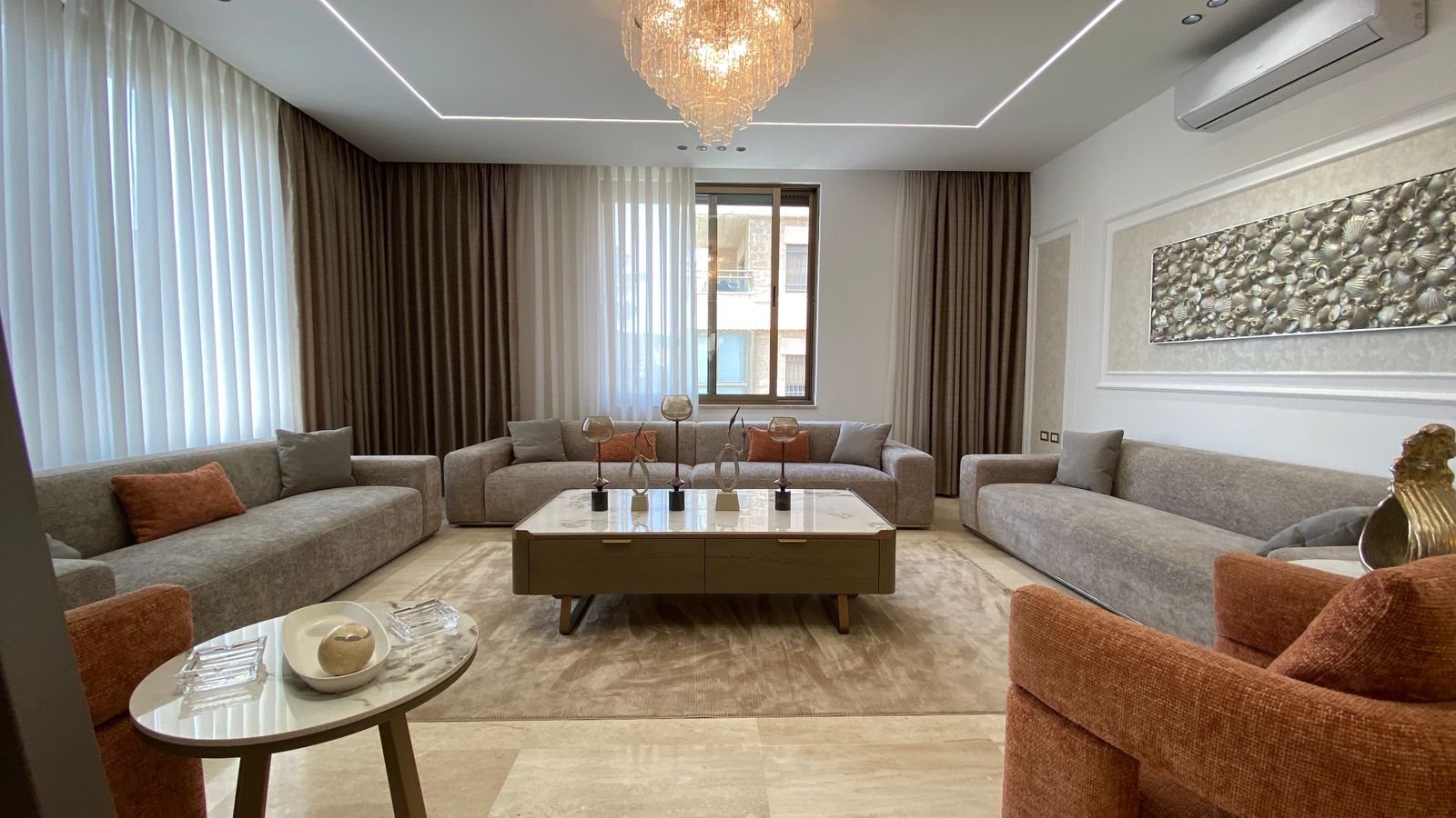 9b22c30a-4447-43ed-8292-bba88324f765 Luxurious living room with three gray sectionals, a marble coffee table, and a crystal chandelier above a neutral rug.