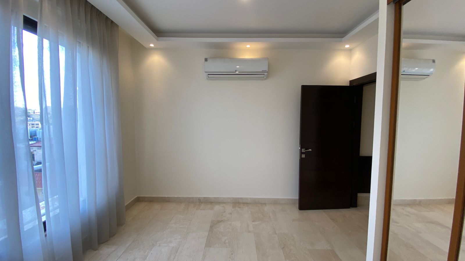 9c23e6df-c932-45ec-912e-070b0b6673fe Empty modern room with beige walls, sheer blue curtains over a large window, a wall-mounted AC unit, and an open dark wooden door.