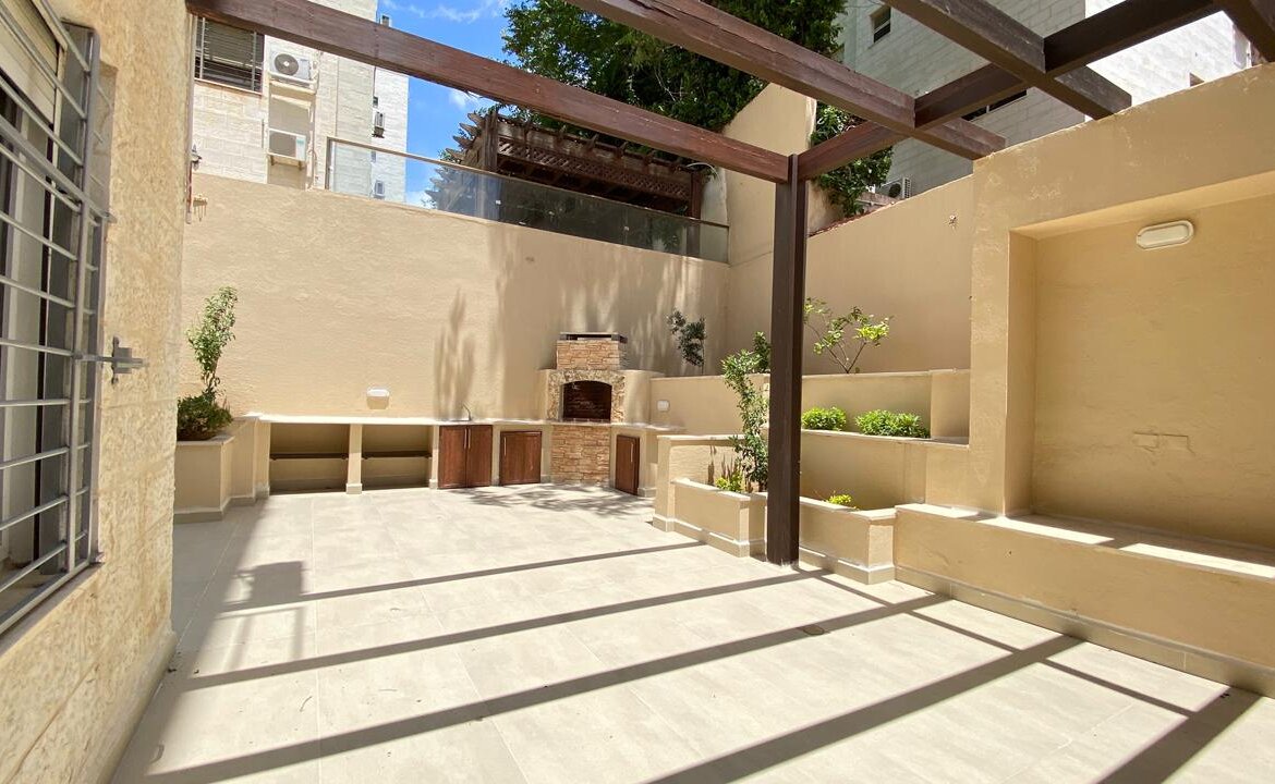 Sunny beige courtyard with a brick outdoor grill, built-in counters and cabinets, and raised planters along stucco walls under a wooden pergola.