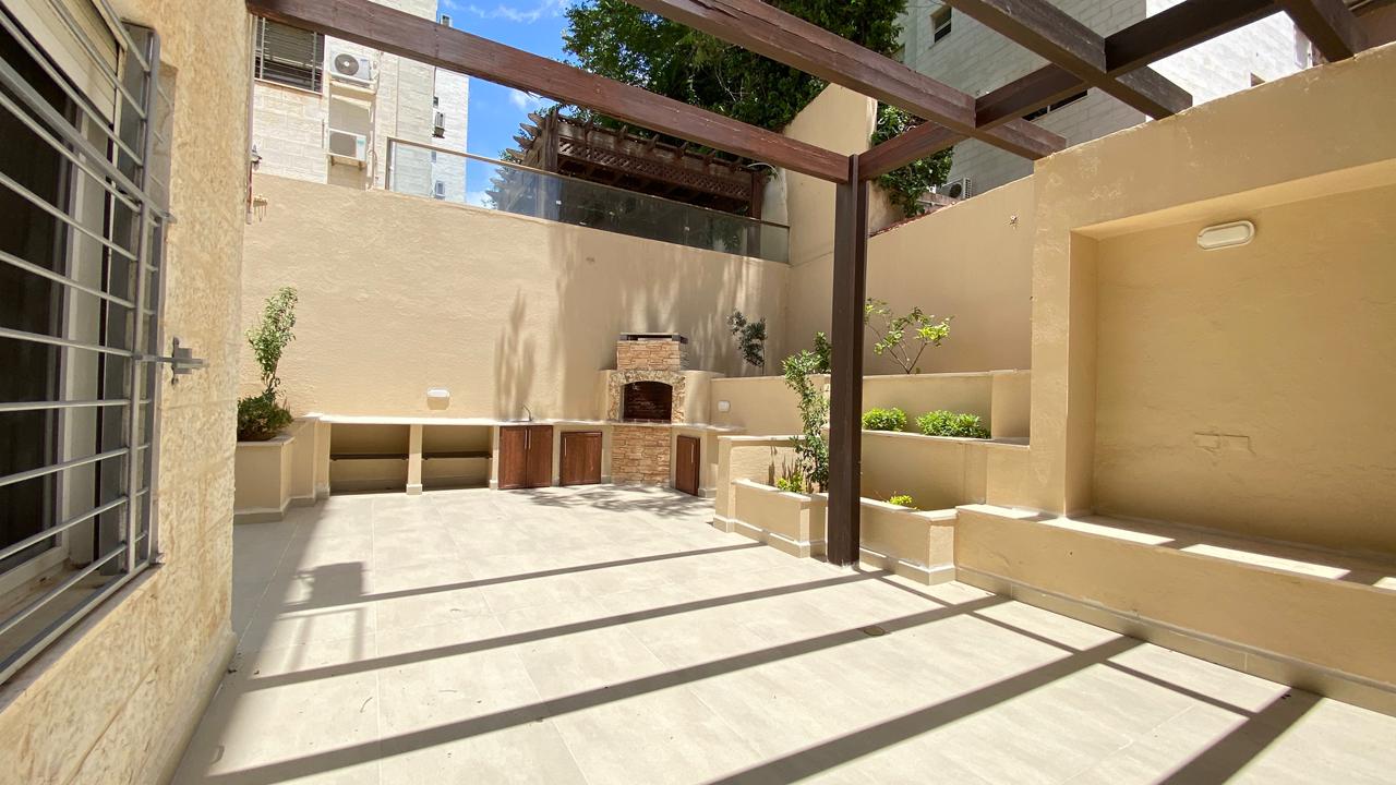 9c3b86d2-759d-483d-865a-348ec01d0f70 Sunny beige courtyard with a brick outdoor grill, built-in counters and cabinets, and raised planters along stucco walls under a wooden pergola.