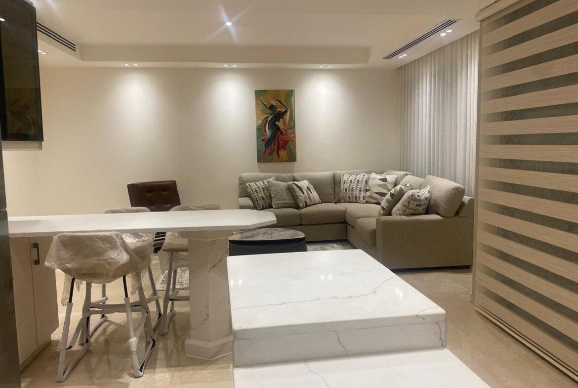 Modern open-plan living space with marble countertops, beige sectional sofa, and a colorful abstract painting above the seating area.