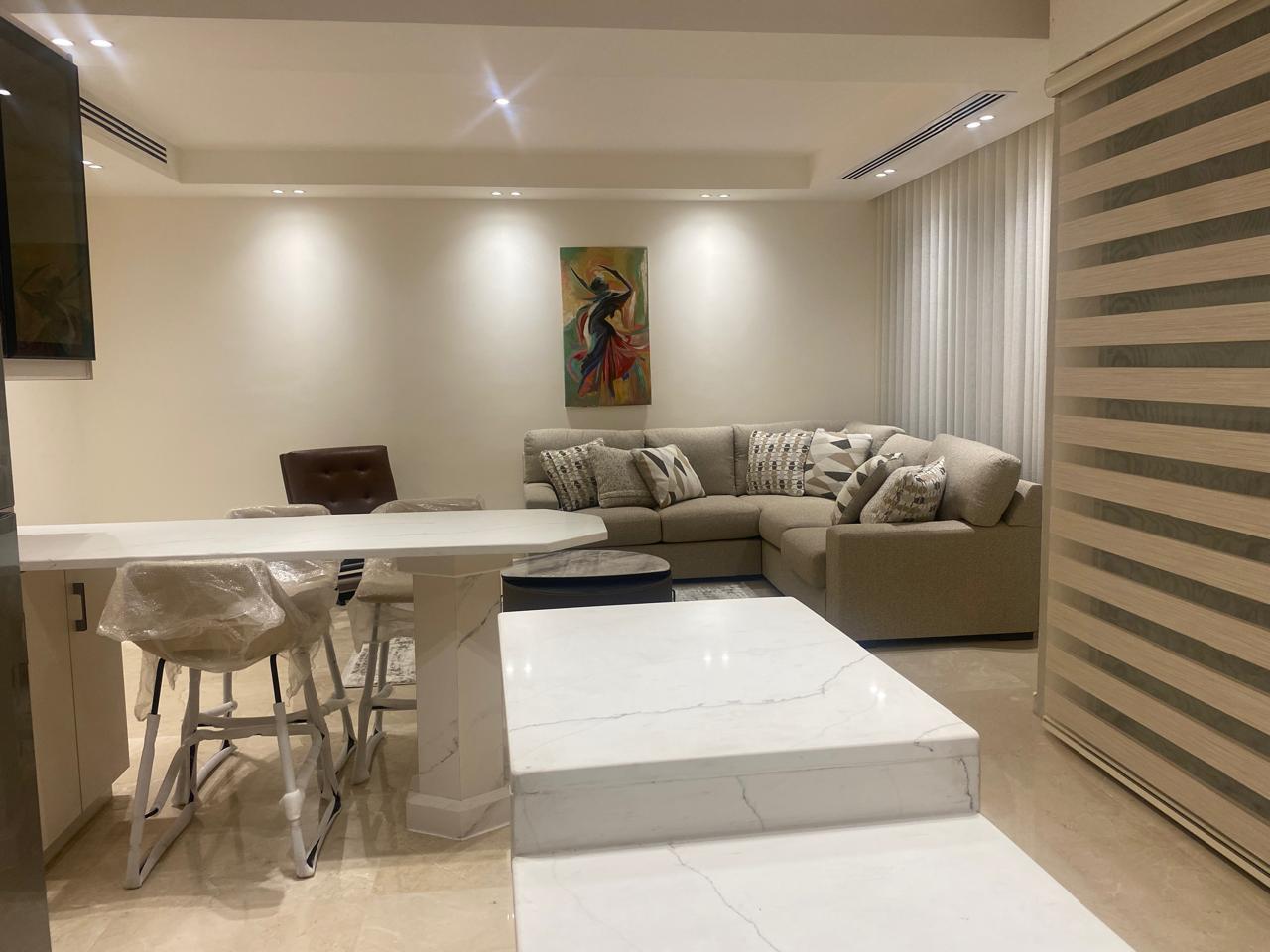 Modern open-plan living space with marble countertops, beige sectional sofa, and a colorful abstract painting above the seating area.