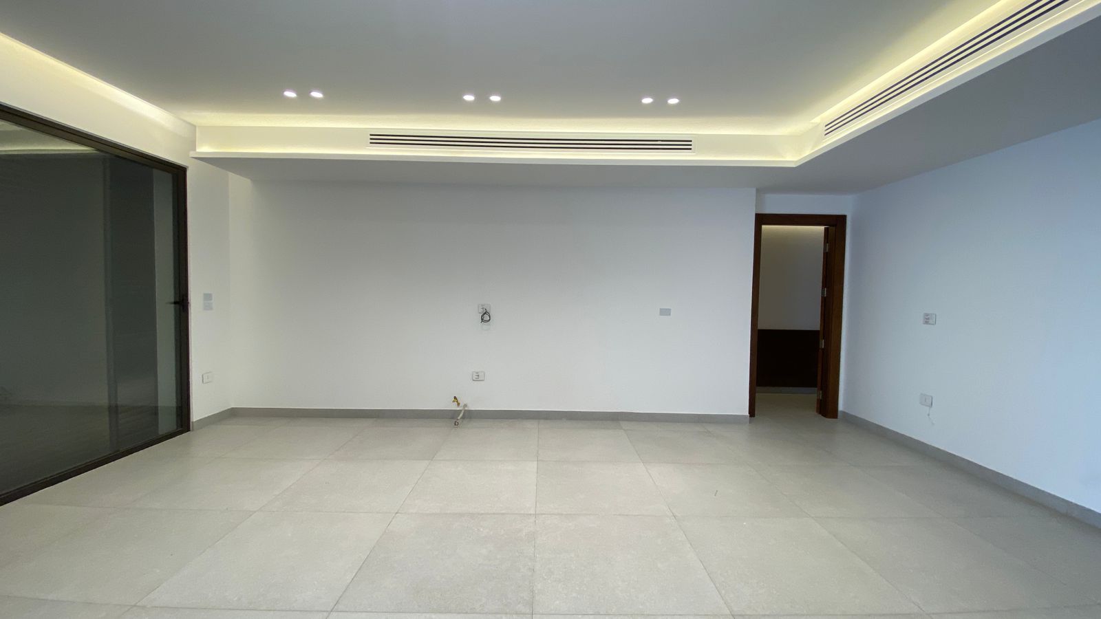 9d23831f-0476-4c06-9e8d-b194b2ebd1aa Empty modern living room with large glass sliding doors on the left, white walls, and recessed ceiling lighting with Cove lights along the ceiling edge.