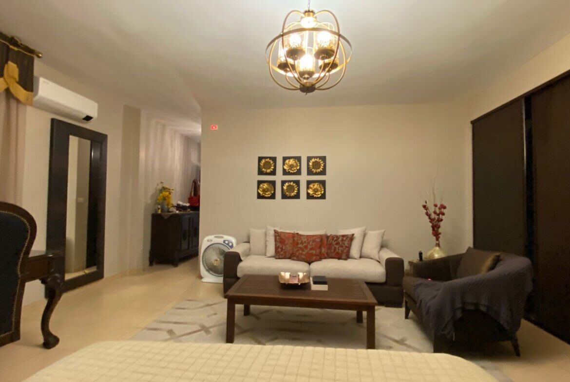 9d4b23d0-6777-43cc-883d-36d577b0d5e1 Living room with a white sofa and red patterned cushions, dark wood coffee table, and a matching dark armchair; wall art of six gold sunflowers above the sofa.