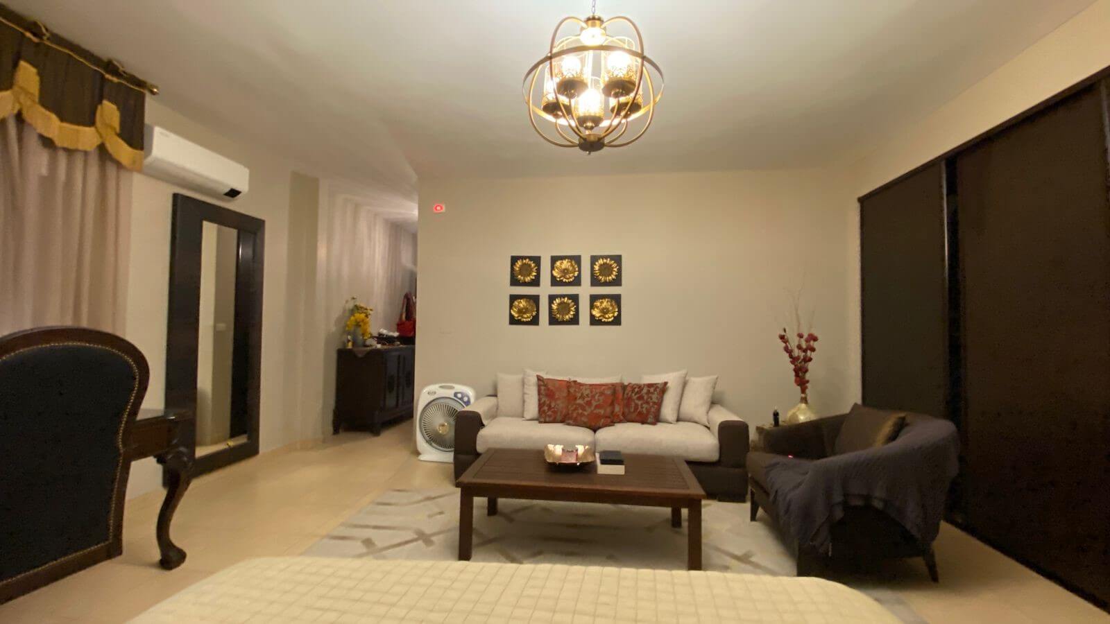 9d4b23d0-6777-43cc-883d-36d577b0d5e1 Living room with a white sofa and red patterned cushions, dark wood coffee table, and a matching dark armchair; wall art of six gold sunflowers above the sofa.