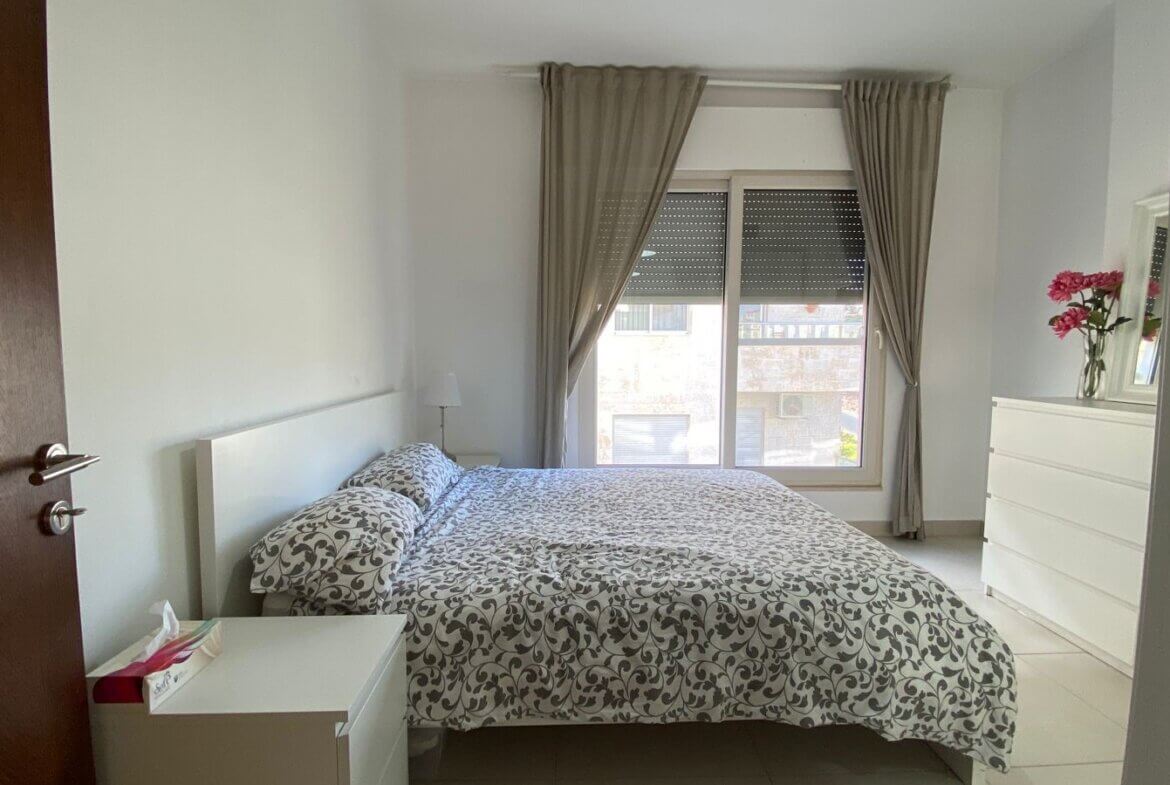 9d51f11e-d21e-4893-8bac-09ada1a77b84 Bright bedroom with a double bed, white furniture, and a large window with beige curtains courtyards light entry point.