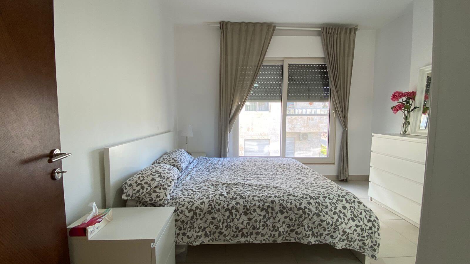 9d51f11e-d21e-4893-8bac-09ada1a77b84 Bright bedroom with a double bed, white furniture, and a large window with beige curtains courtyards light entry point.
