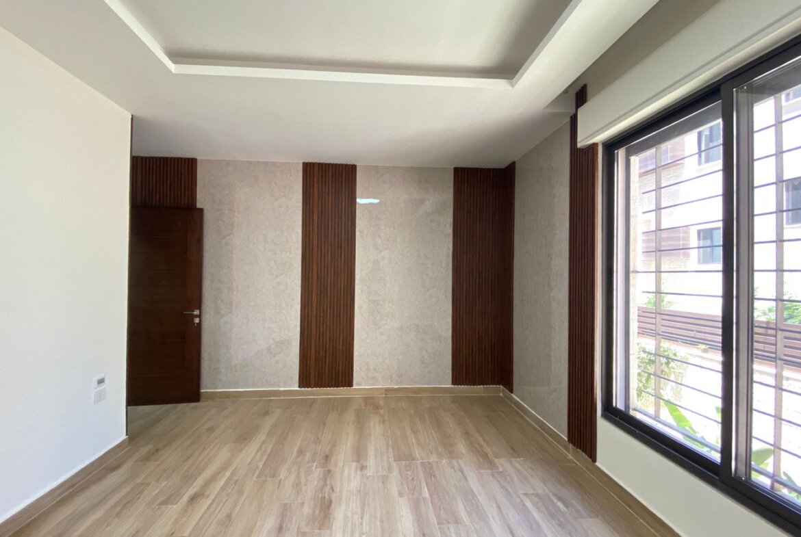 Modern empty living room with light wood flooring, beige walls, and vertical dark wood panels; large window with metal bars on the right and a wooden door on the back wall.