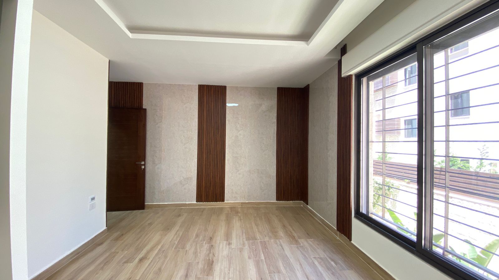 9d5c7f31-46e3-45d9-acc3-1fe226b8c523 Modern empty living room with light wood flooring, beige walls, and vertical dark wood panels; large window with metal bars on the right and a wooden door on the back wall.