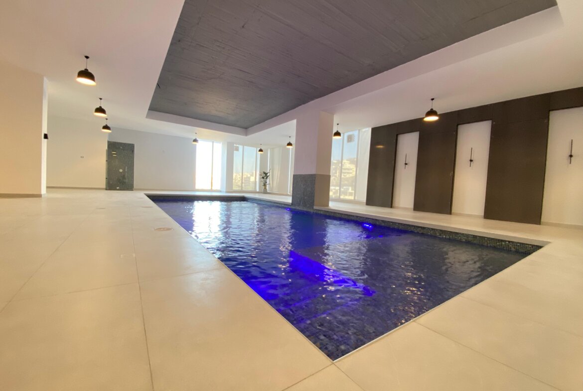 Indoor pool area with blue water, white tiles, and a dark curved ceiling illuminated by pendant lights