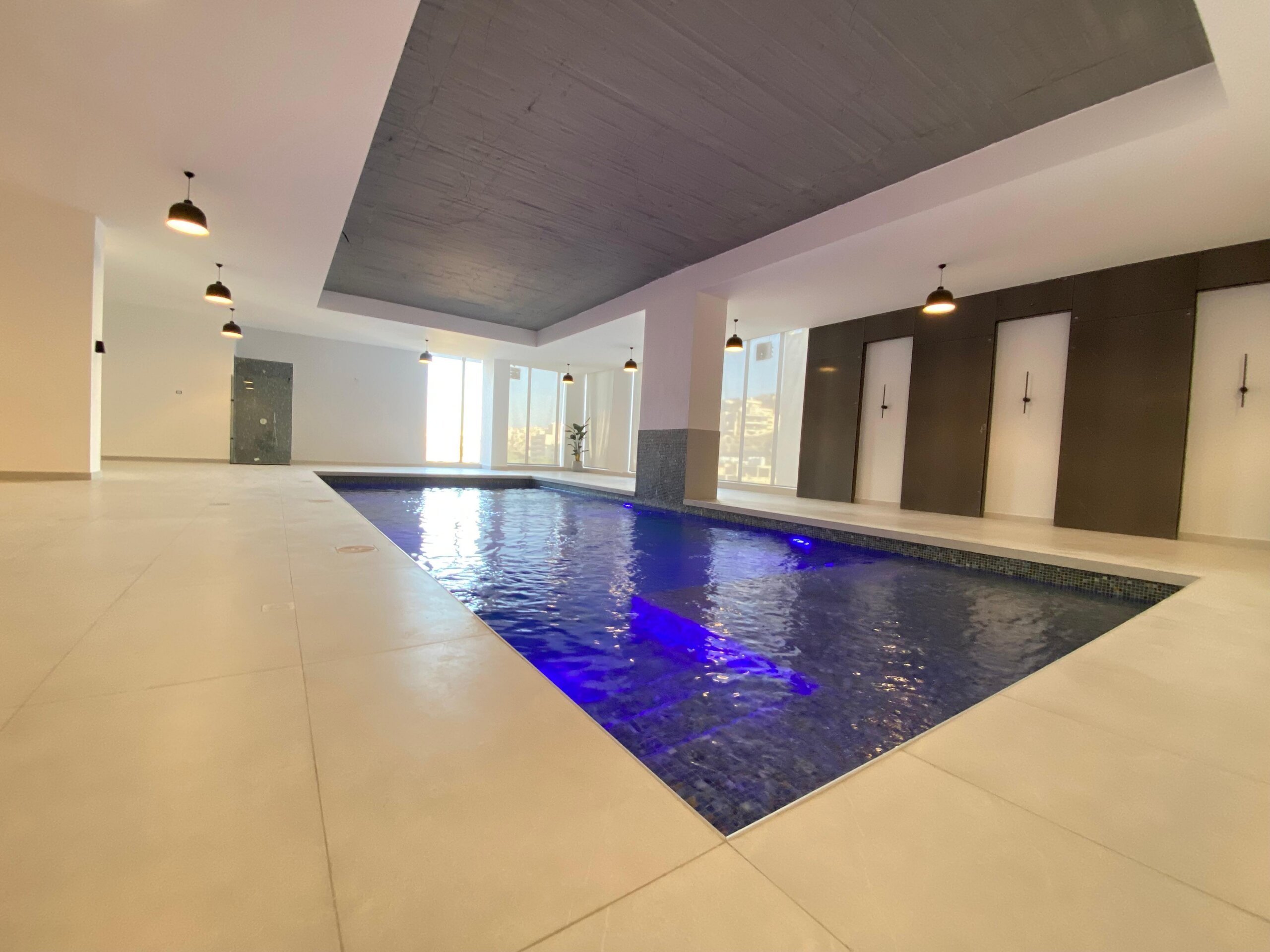9d6aadce-aba6-4a2e-8bfb-5245063cfda6 Indoor pool area with blue water, white tiles, and a dark curved ceiling illuminated by pendant lights