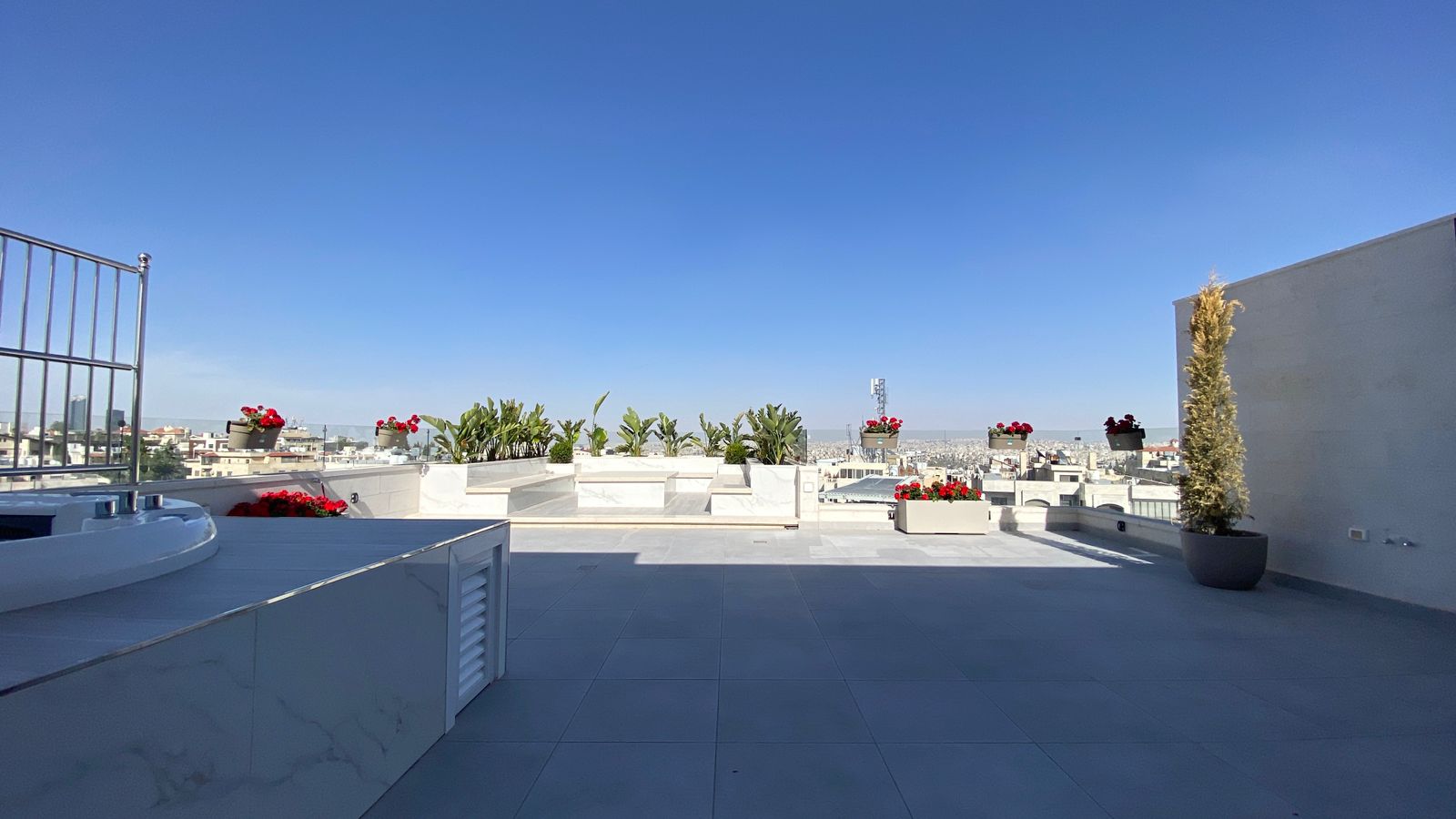 9d85d582-2fe2-4e37-84b3-87d119518b16 Open rooftop terrace with white marble planters, red flowers, and potted greenery overlooking a cityscape under a clear blue sky.