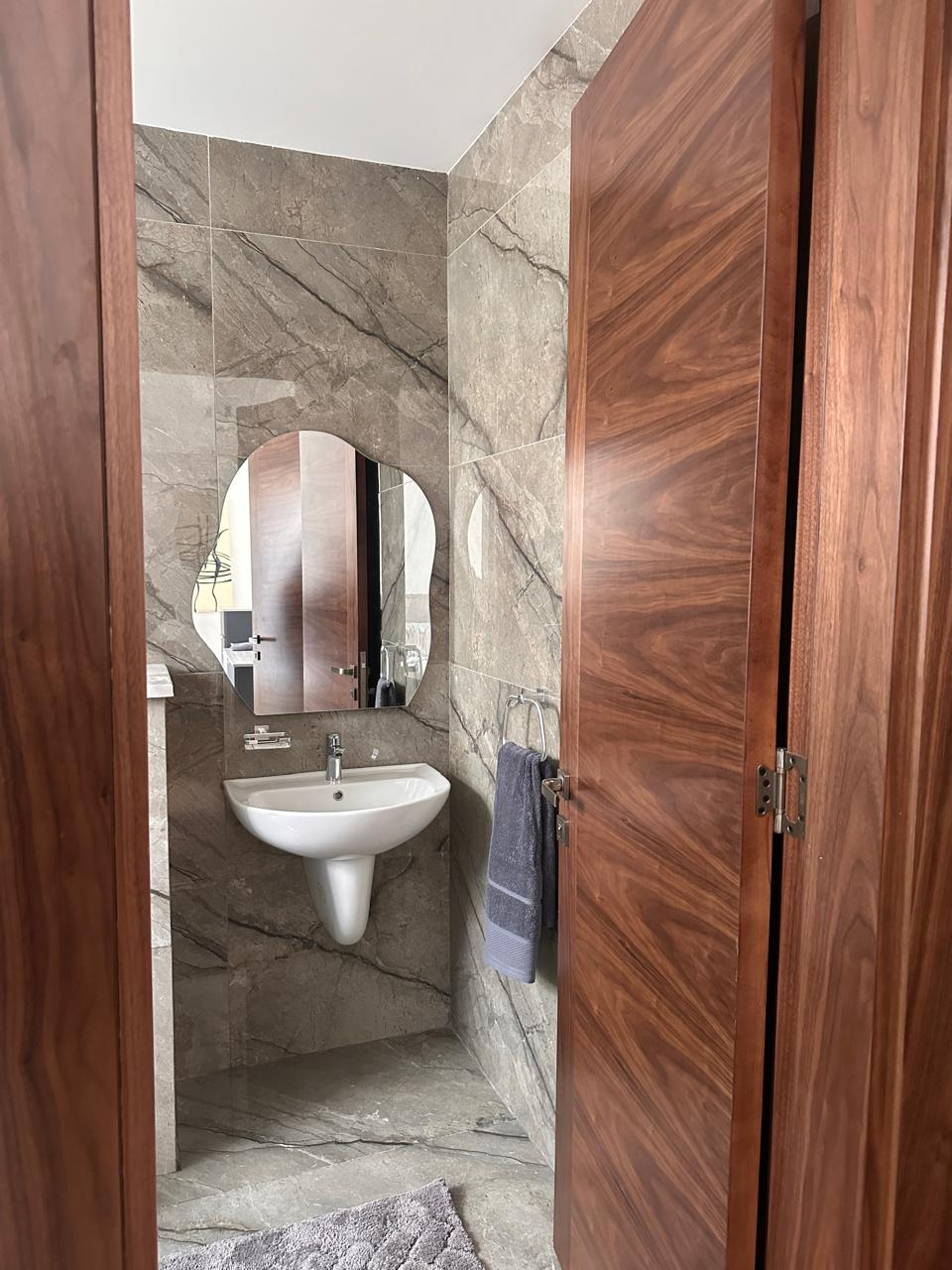9df93797-6212-4bf2-ba67-dda10db11f2e Small bathroom with gray marble walls, a white pedestal sink, oval mirror, and wooden doors.