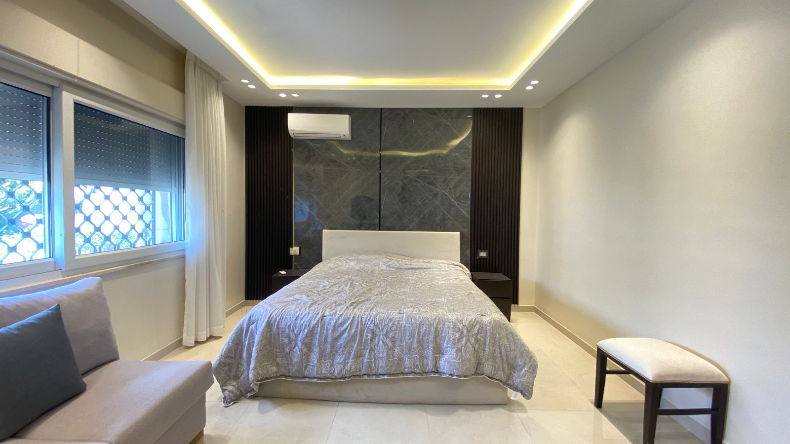 9e2910e7-d8d7-4b16-9731-418cdcf8b3e8 Modern bedroom with a king bed, white headboard, and gray patterned bedding against a dark marble headboard panel. A ceiling cove light glows above.