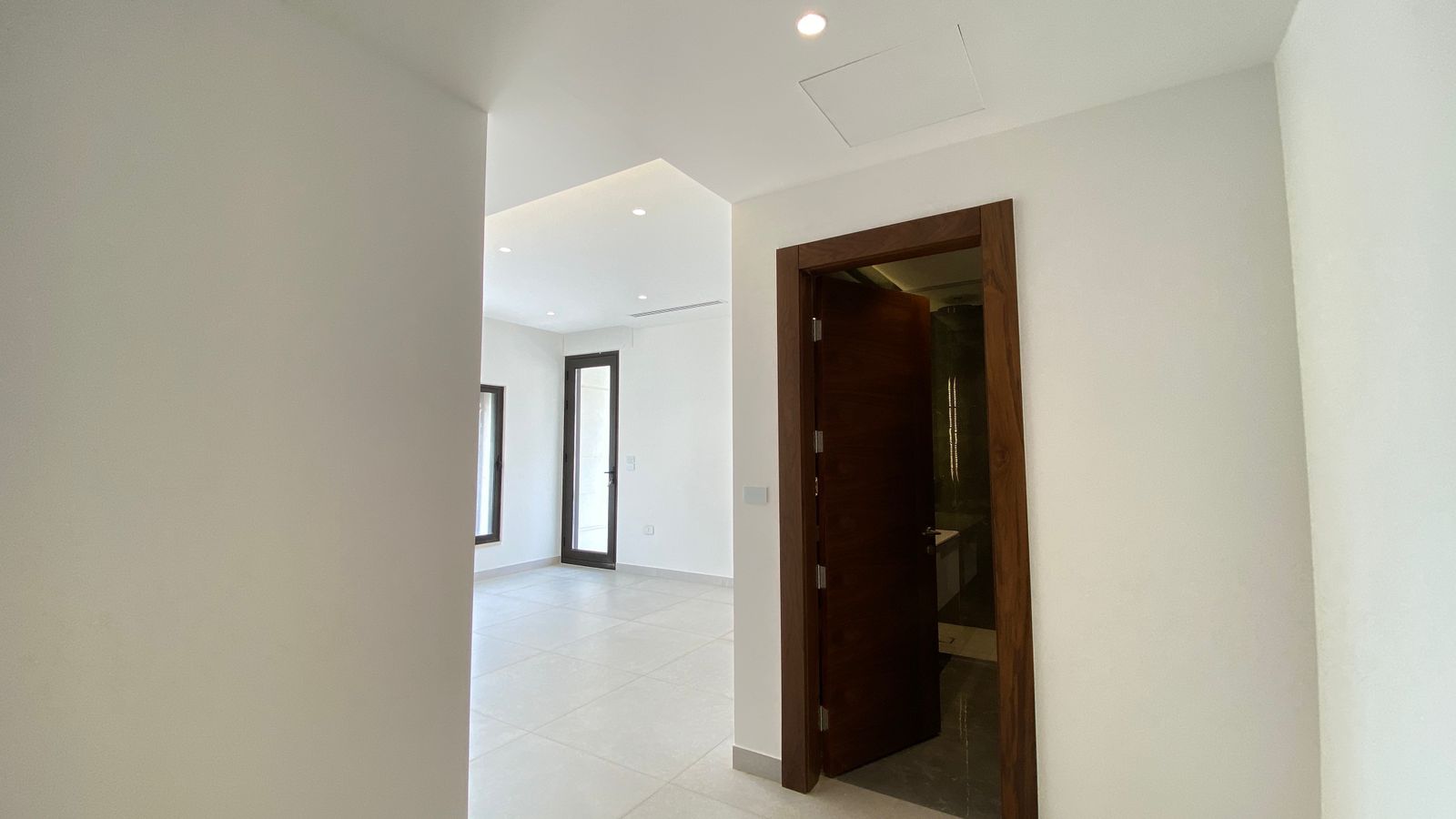 9e38a0e6-bfdc-4065-8a06-2d5b2c730516 Bright, modern interior: white walls and tiled floor, a dark wood doorway opens into a bathroom, with natural light coming from the adjacent room.