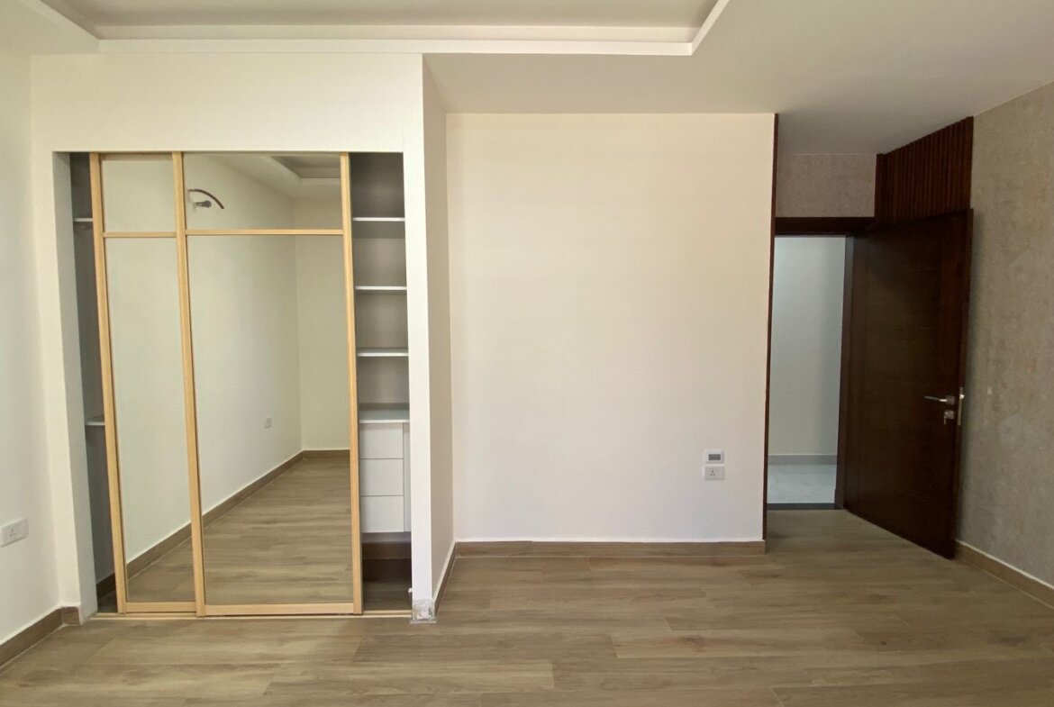 Bedroom with light wood floor, built-in wardrobe with sliding mirrored doors on the left, and a dark wooden door open to another room on the right.