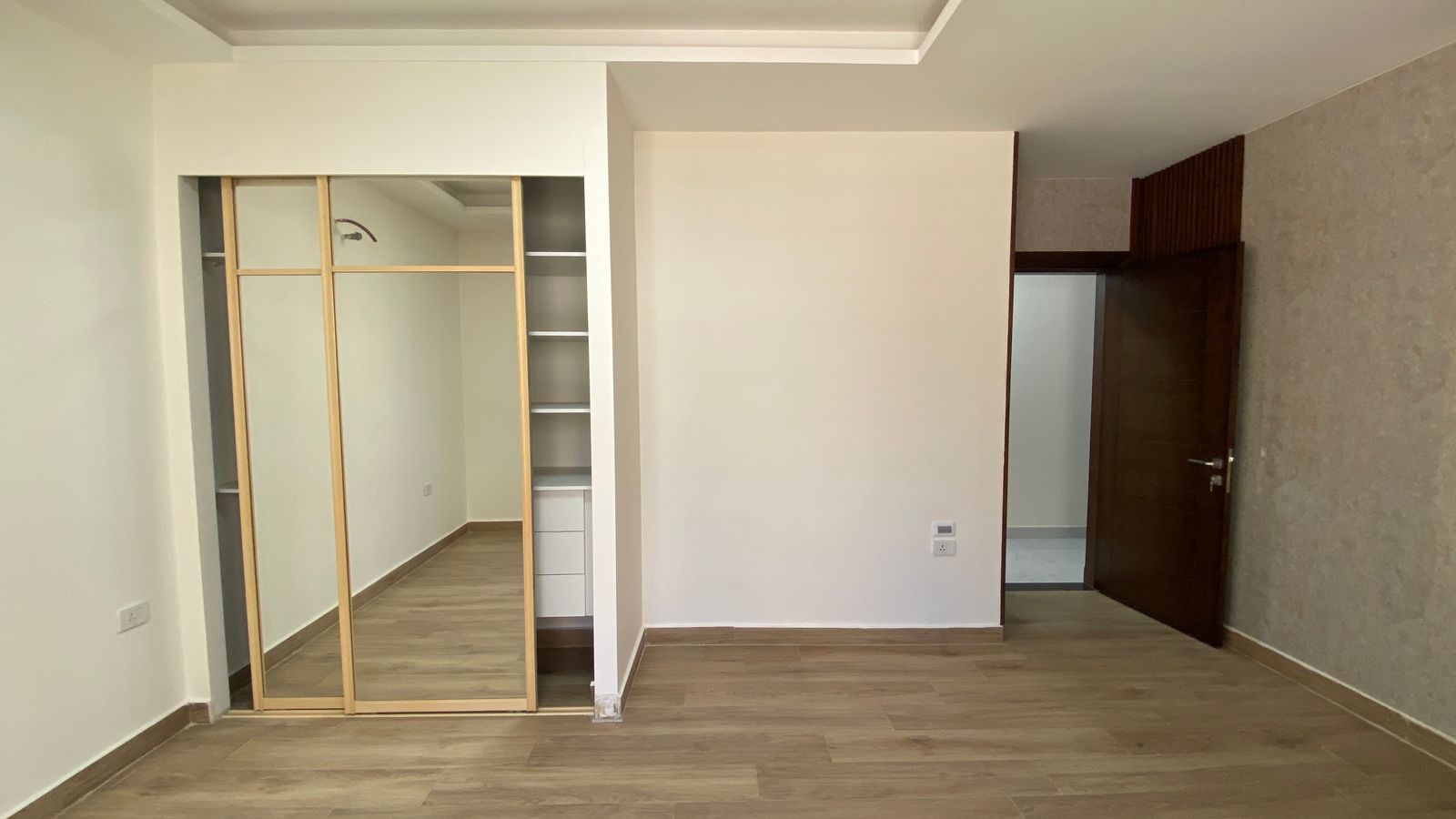 9eb8fa09-5521-48f5-a07a-cc0558edc953 Bedroom with light wood floor, built-in wardrobe with sliding mirrored doors on the left, and a dark wooden door open to another room on the right.