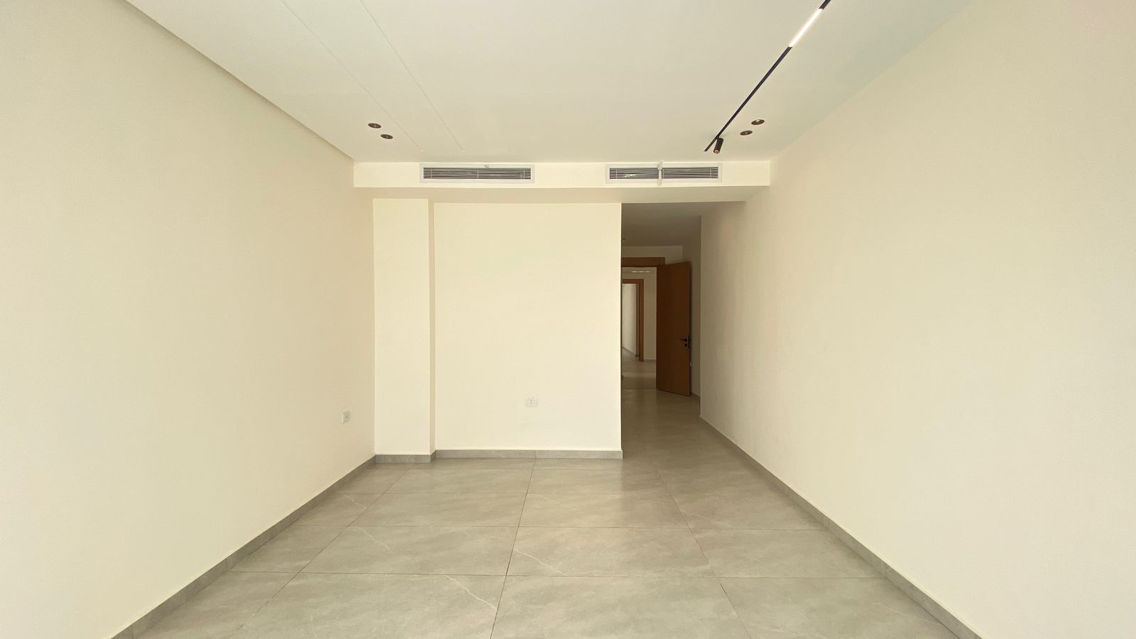 9f184e46-f7c2-4c06-b4d7-3c12b1dc7998 Empty beige-walled hallway with a gray tiled floor and an open door at the far end.