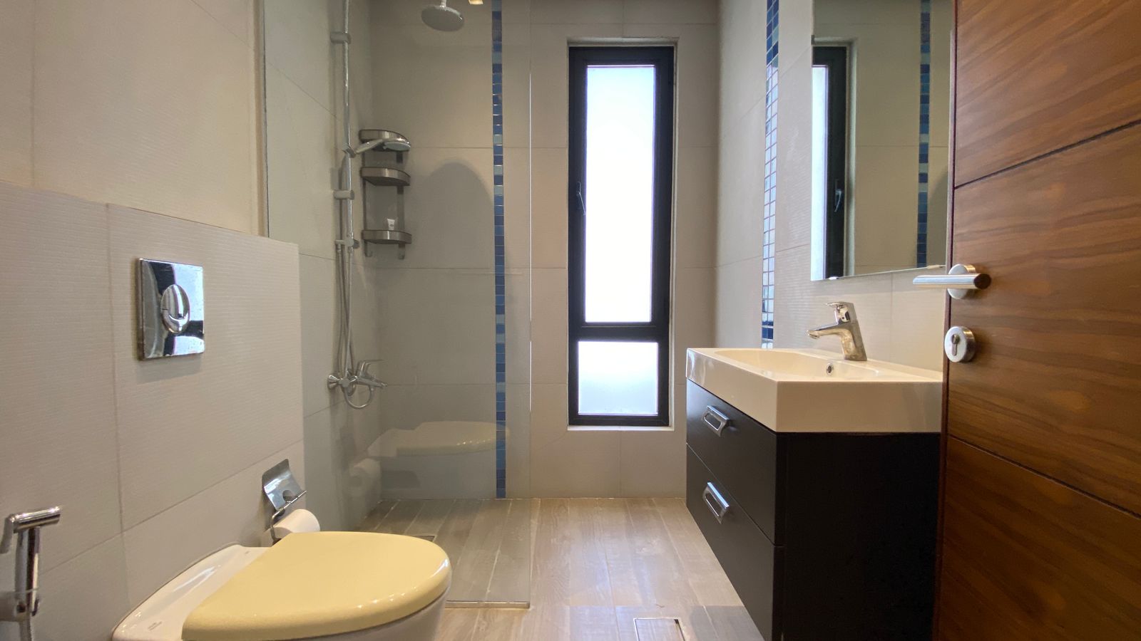 9f734ff4-296e-46c3-a90e-b84efeef8c31 Modern bathroom with glass-walled shower, toilet, and a white sink on a dark vanity under a tall narrow window. A wooden door is ajar.