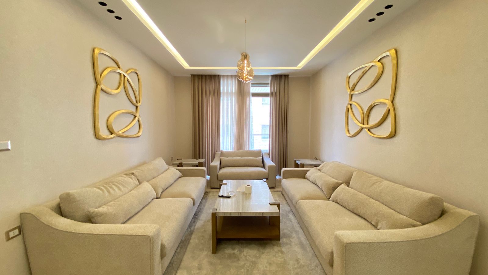 9fcc922c-4b3b-494e-bc49-c9c30dae40ec Beige living room with two sofas and a matching loveseat around a central wooden coffee table, with gold abstract wall sculptures on each side.