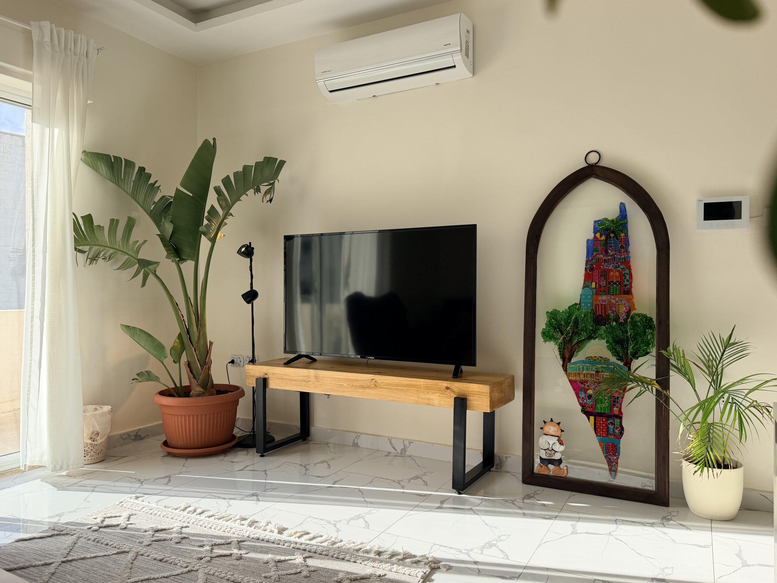 IMG_2124 copy Living room corner with a wall-mounted white air conditioner above a TV on a wooden stand, flanked by tall potted plants and a colorful arch-framed mural.
