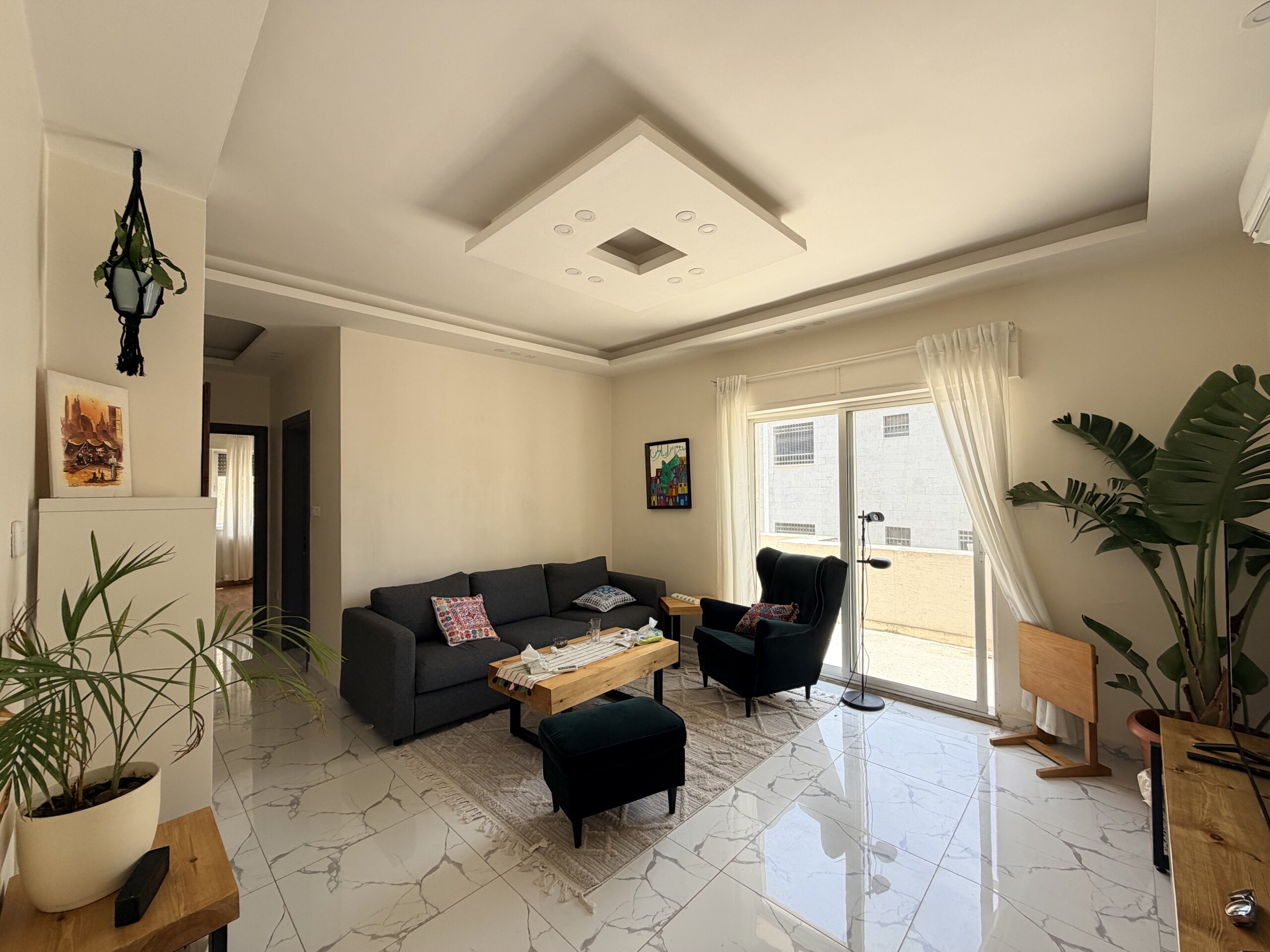 IMG_2893 Bright living room with marble-tiled floor, gray sofa, wooden coffee table, and large windows with sheer white curtains; several potted plants nearby.
