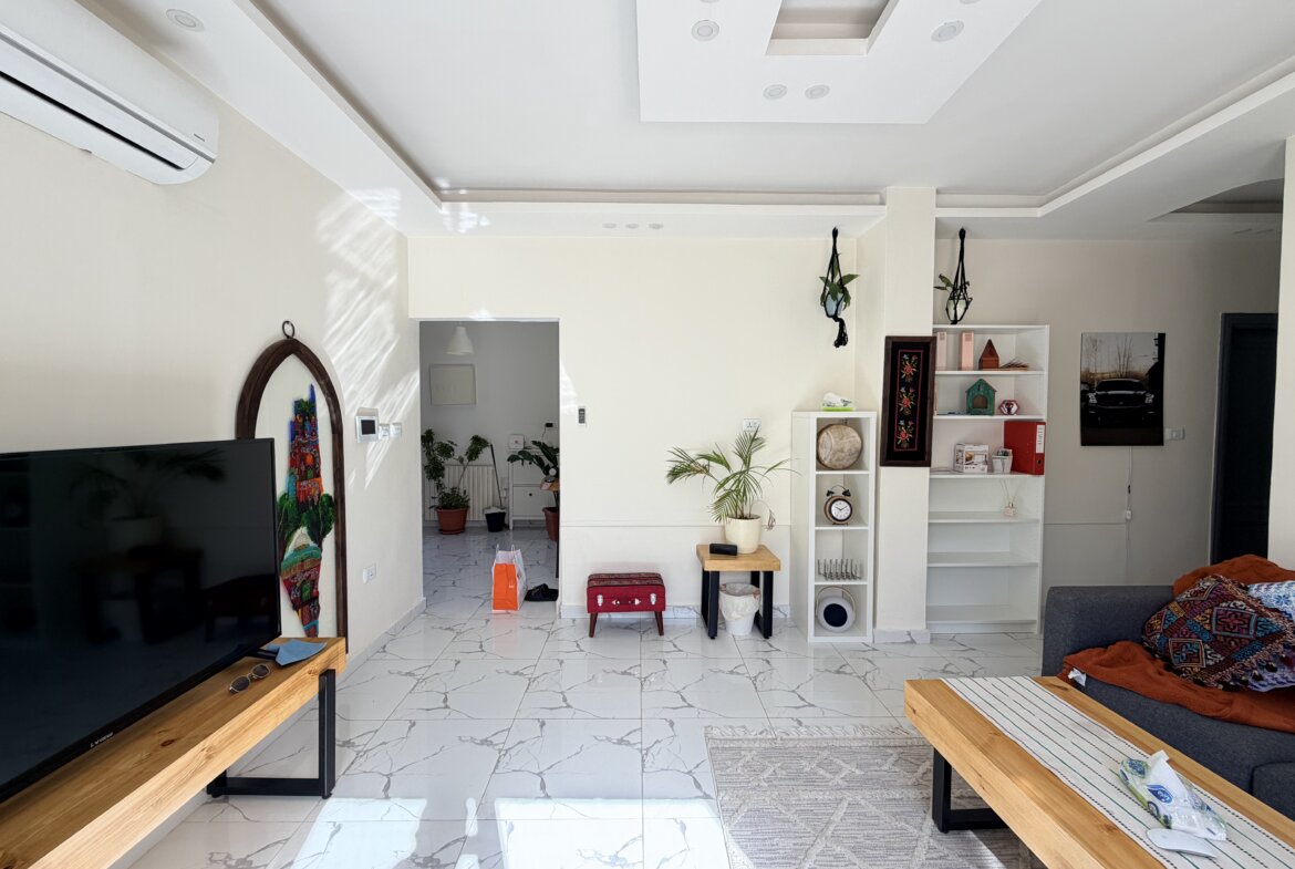 Bright open living room with a wall-mounted flat-screen TV on the left, two wooden benches, and white marble flooring with gray veining.
