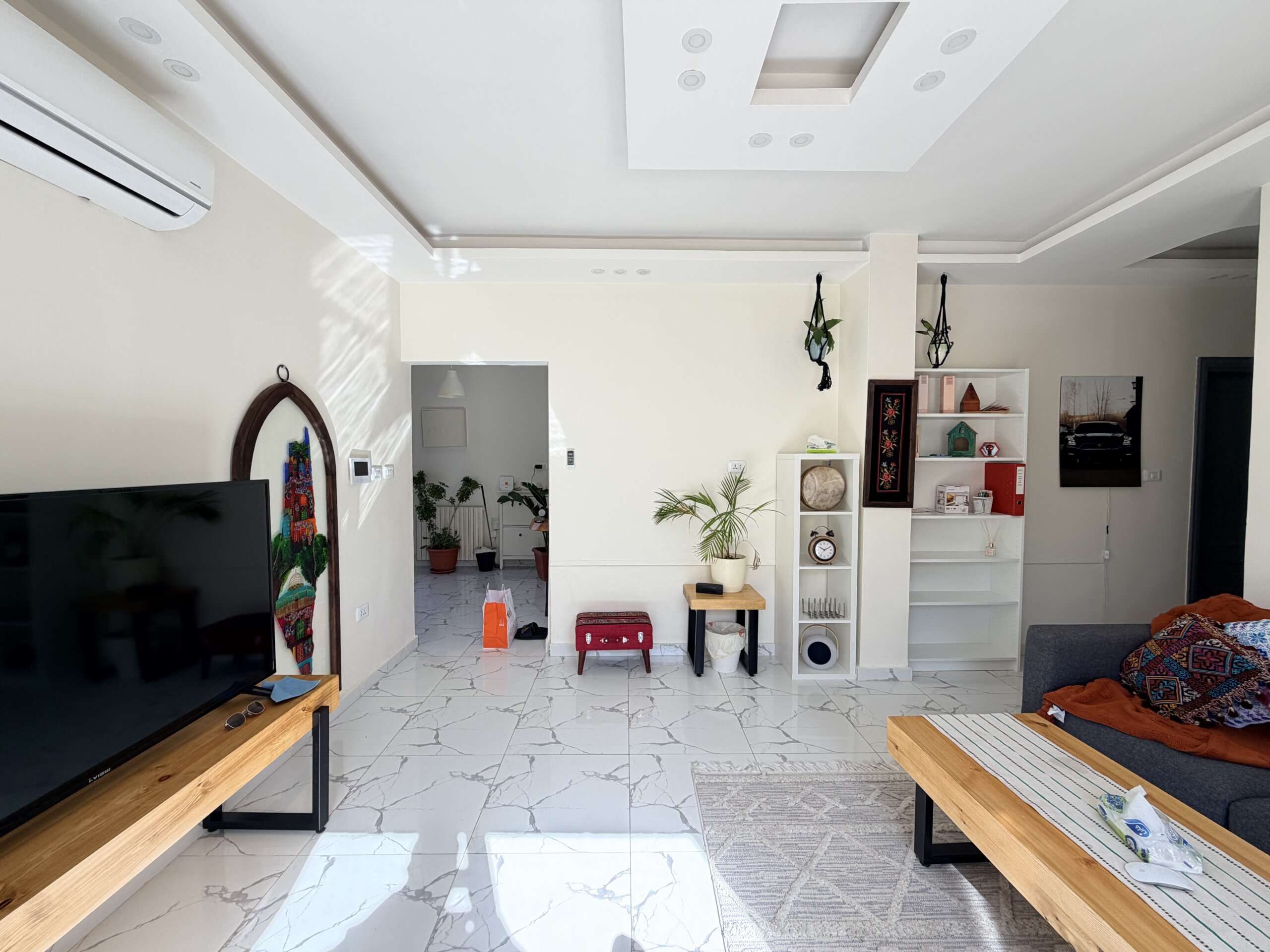 IMG_2945 Bright open living room with a wall-mounted flat-screen TV on the left, two wooden benches, and white marble flooring with gray veining.