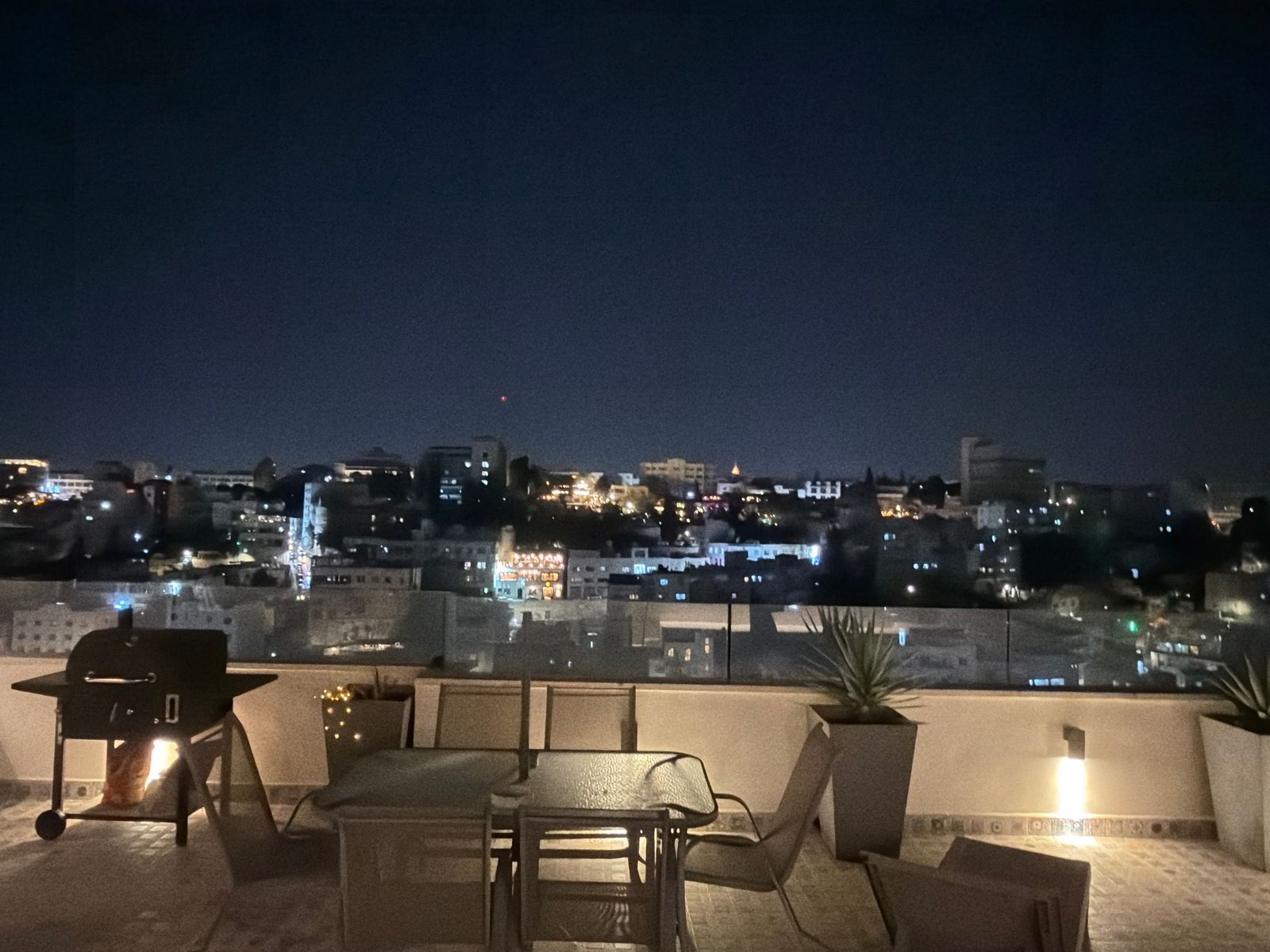 WhatsApp Image 2026-05-07 at 84327 PM 1 Night rooftop terrace with seating and a grill, overlooking a lit city skyline under a dark sky.