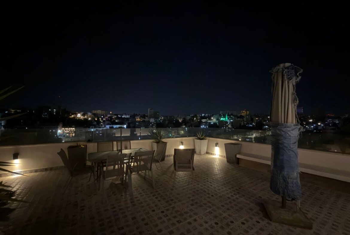 Rooftop terrace at night with a table, chairs, and city lights in the distance.