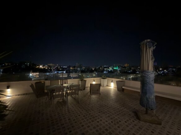 Rooftop terrace at night with a table, chairs, and city lights in the distance.