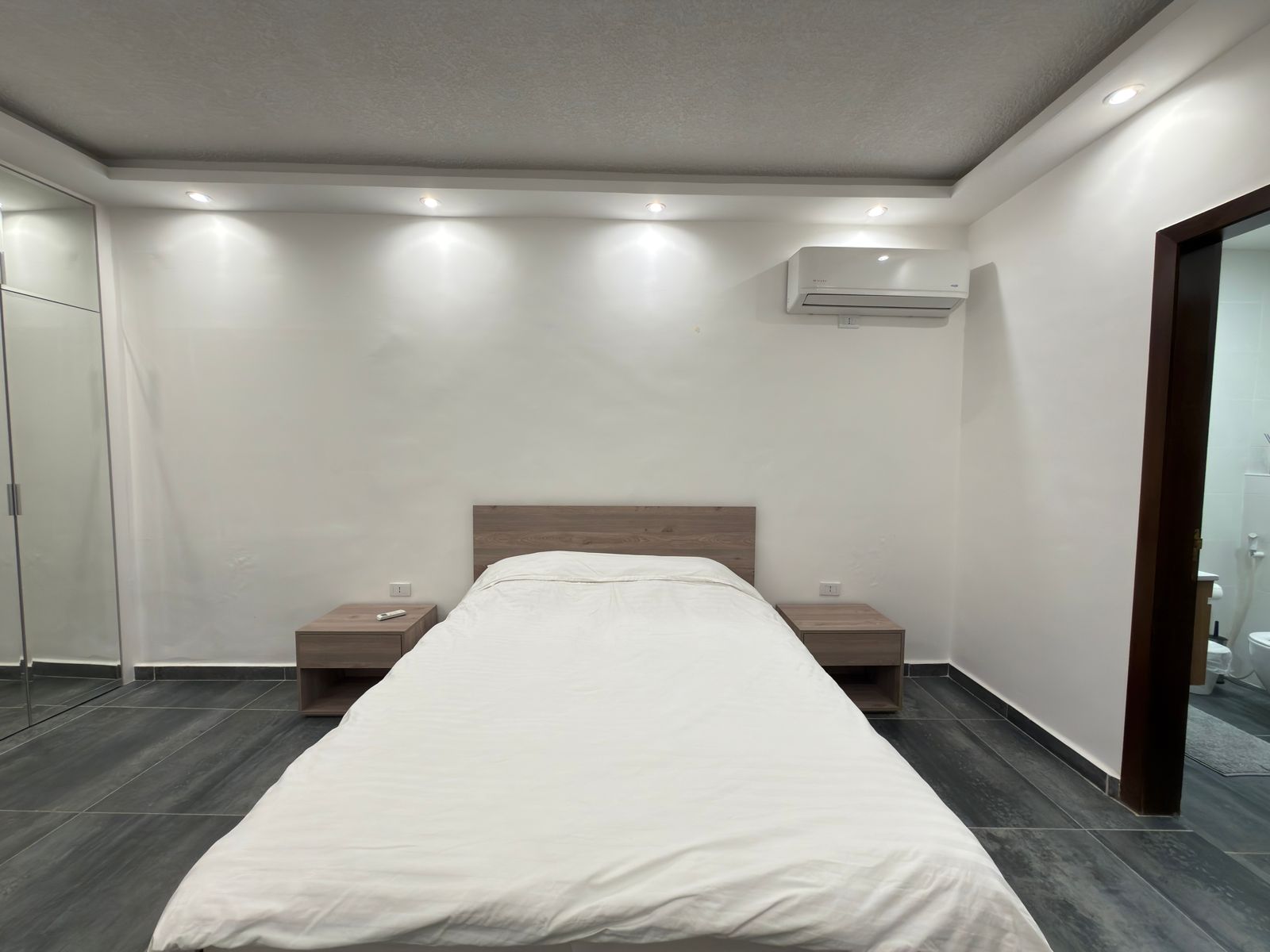 WhatsApp Image 2026-05-07 at 84331 PM 1 Minimalist bedroom with a double bed, wood headboard, two nightstands, white walls, and recessed ceiling lights.