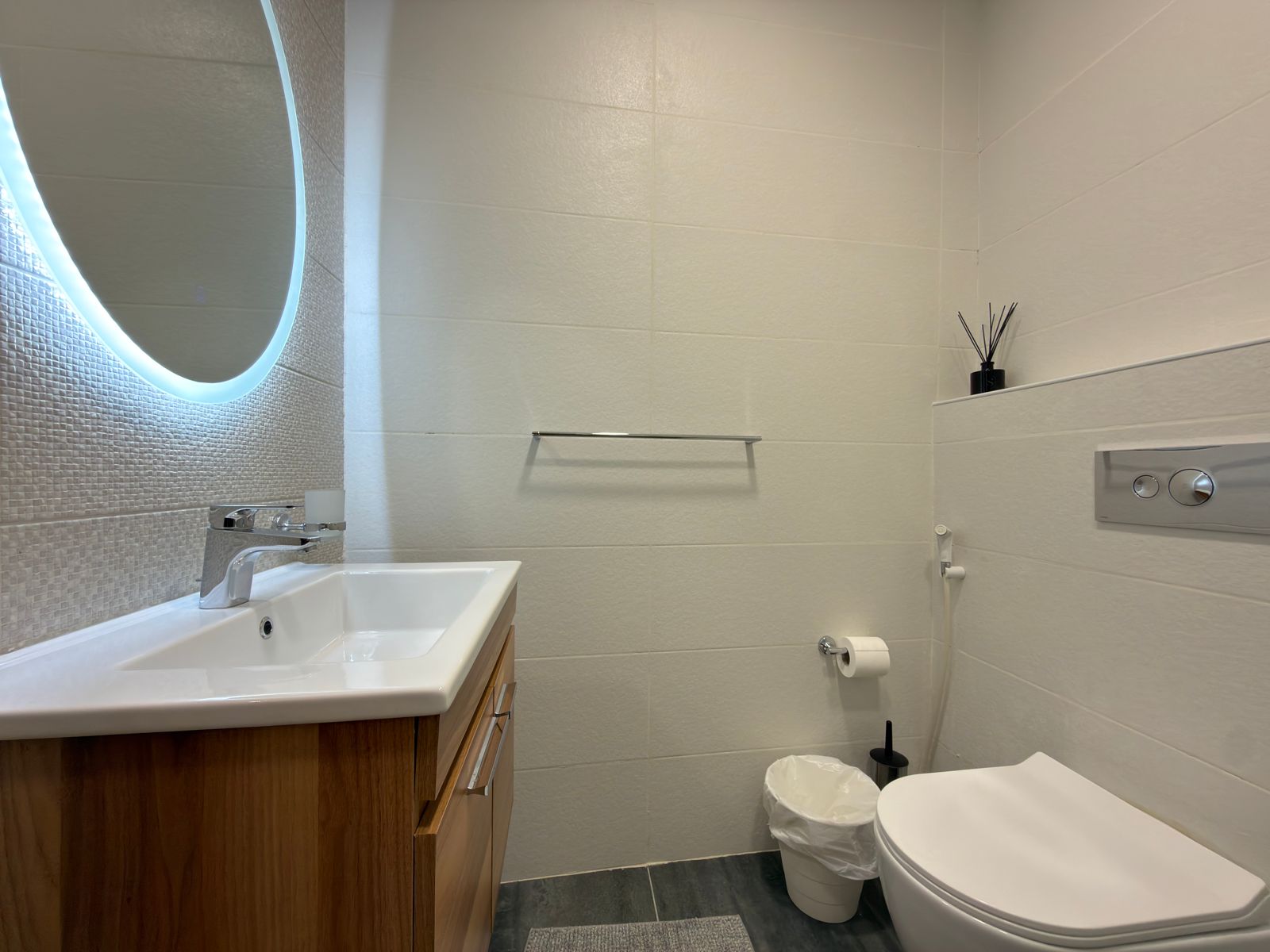 WhatsApp Image 2026-05-07 at 84331 PM Round backlit mirror over a white rectangular sink with a wooden vanity in a modern bathroom setting.