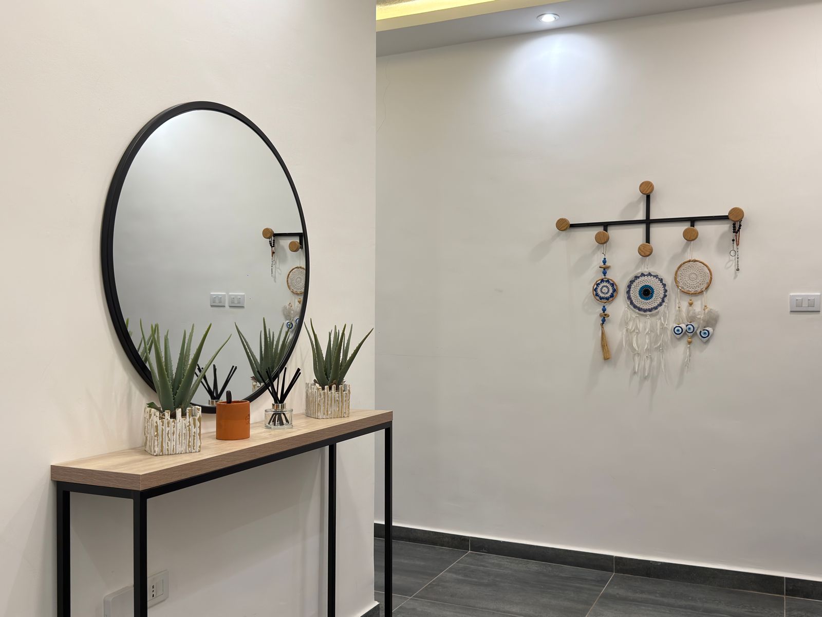 WhatsApp Image 2026-05-07 at 84332 PM 2 Round black-framed mirror above a wooden console table with green succulents and reed diffusers; wall display of blue-and-white dreamcatchers on a horizontal rack to the right.