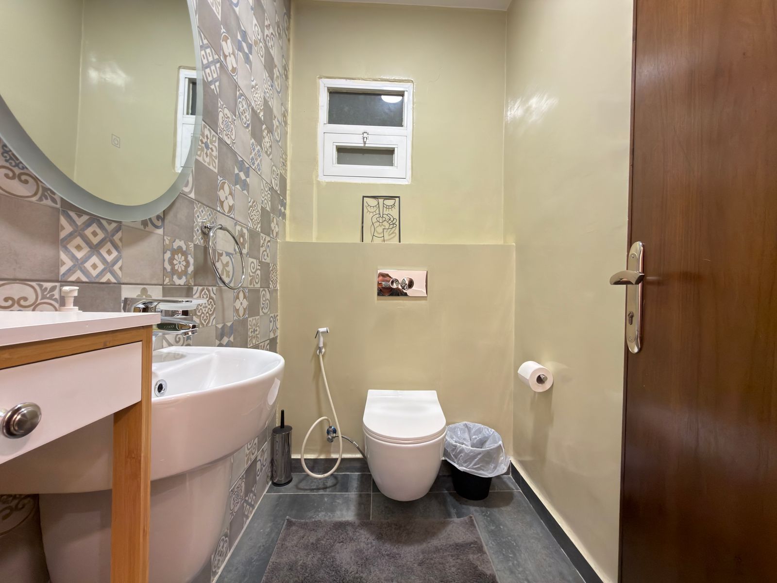 WhatsApp Image 2026-05-07 at 84332 PM 3 Small bathroom with a round mirror, white sink, and wall-mounted toilet against a patterned tile wall.