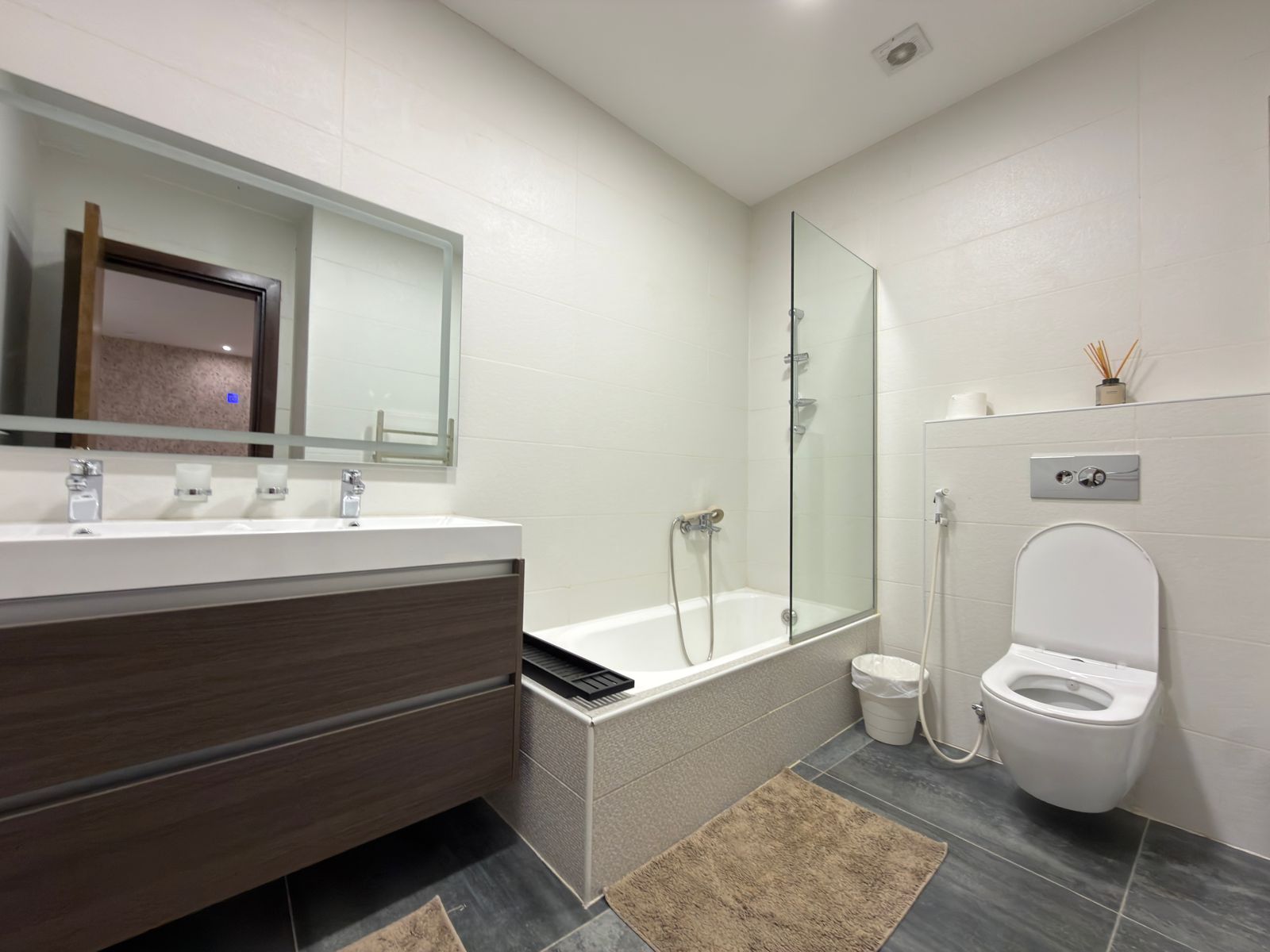 WhatsApp Image 2026-05-07 at 84332 PM Modern bathroom with a double-sink vanity, large mirror, and bathtub with glass shower panel. A toilet sits to the right with a bidet hose and trash can nearby.