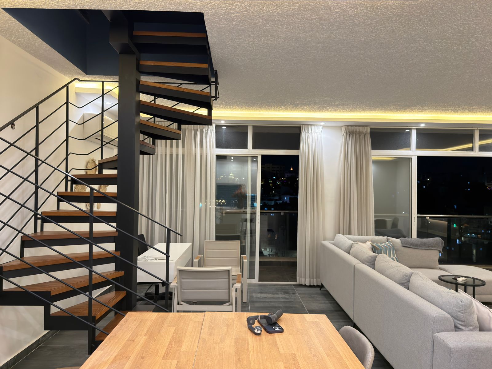 WhatsApp Image 2026-05-07 at 84333 PM Modern living room with a black spiral staircase, gray sectional sofa, and city lights visible through large windows at night.