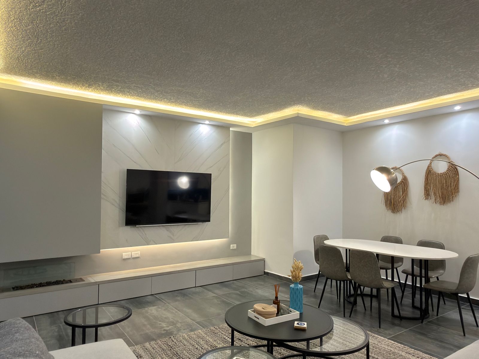 WhatsApp Image 2026-05-07 at 84334 PM 3 Modern living/dining room with a wall-mounted TV on a marble-pattern feature wall and soft under-cabinet lighting.