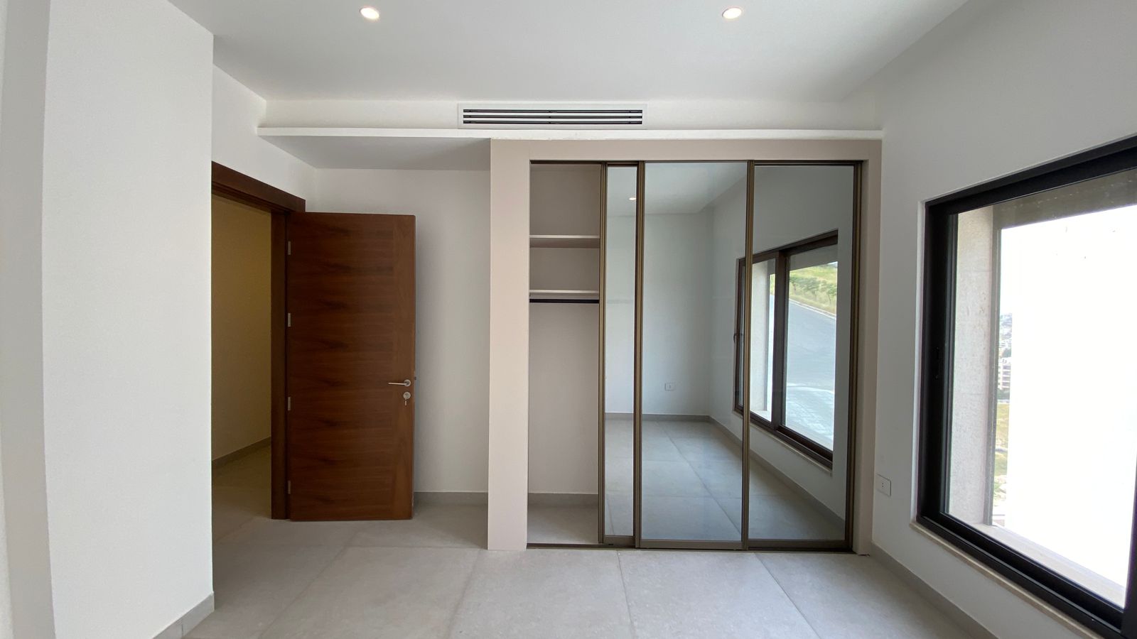 a0190659-1800-4f26-8827-39c743a42f16 Empty modern room with a wooden door ajar, built-in wardrobe with mirrored sliding doors, and large windows revealing an outdoor view