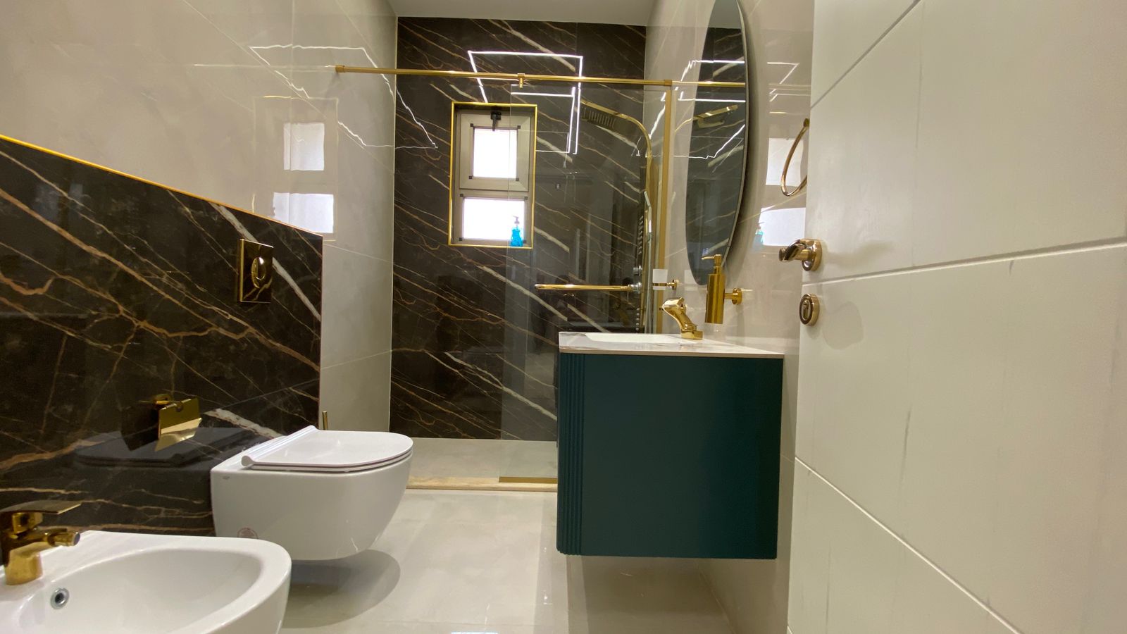 a143278a-dfb9-4ff2-8085-565aa27b8f68 Modern bathroom with black marble walls, white fixtures, and gold hardware including faucet, handles, and towel bar; green vanity under white countertop.