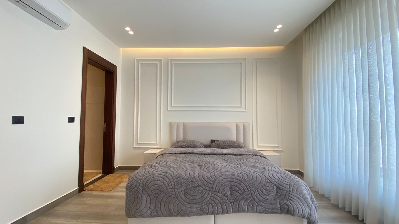 a1596b2a-8fe7-446e-8ddd-97938977f83b Modern bedroom with a low, upholstered bed and gray patterned quilt, white paneled walls, and sheer curtains along the right window wall.