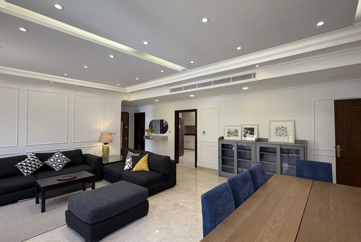 Open-plan living room with dark sectional sofas, a coffee table, and a wooden dining table with blue chairs nearby.