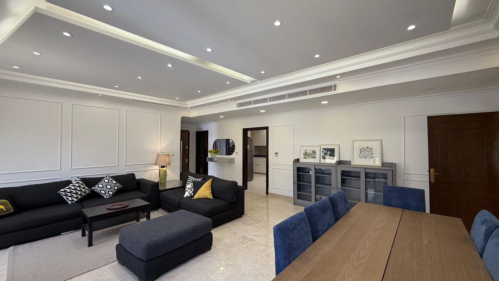Open-plan living room with dark sectional sofas, a coffee table, and a wooden dining table with blue chairs nearby.