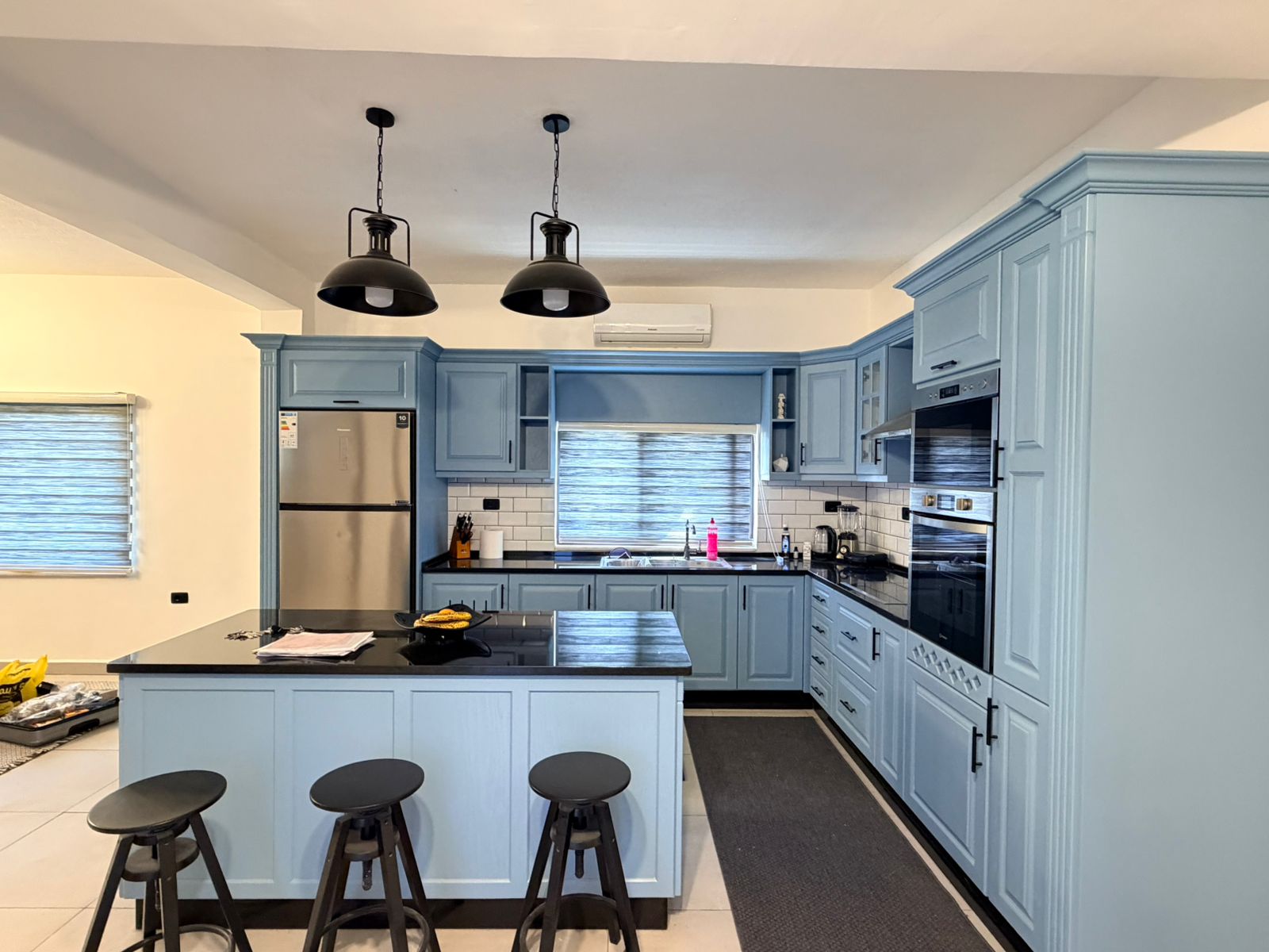 a266dcb0-f956-42cd-a375-8486255ab495 Blue kitchen with an island, black countertops, and two black pendant lights; stainless steel fridge and double wall ovens on the right.