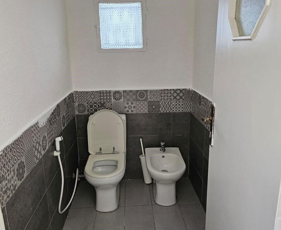 a2f4c815-cb54-41c3-8524-5c70263f2b11 Small bathroom with a white toilet and a nearby bidet, handheld spray, and gray tile walls with decorative pattern mid-height and a small frosted window.