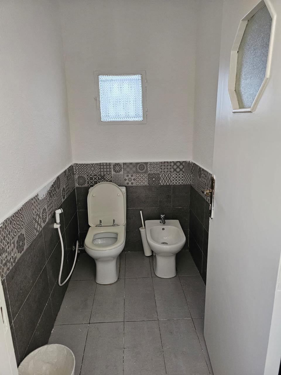 a2f4c815-cb54-41c3-8524-5c70263f2b11 Small bathroom with a white toilet and a nearby bidet, handheld spray, and gray tile walls with decorative pattern mid-height and a small frosted window.