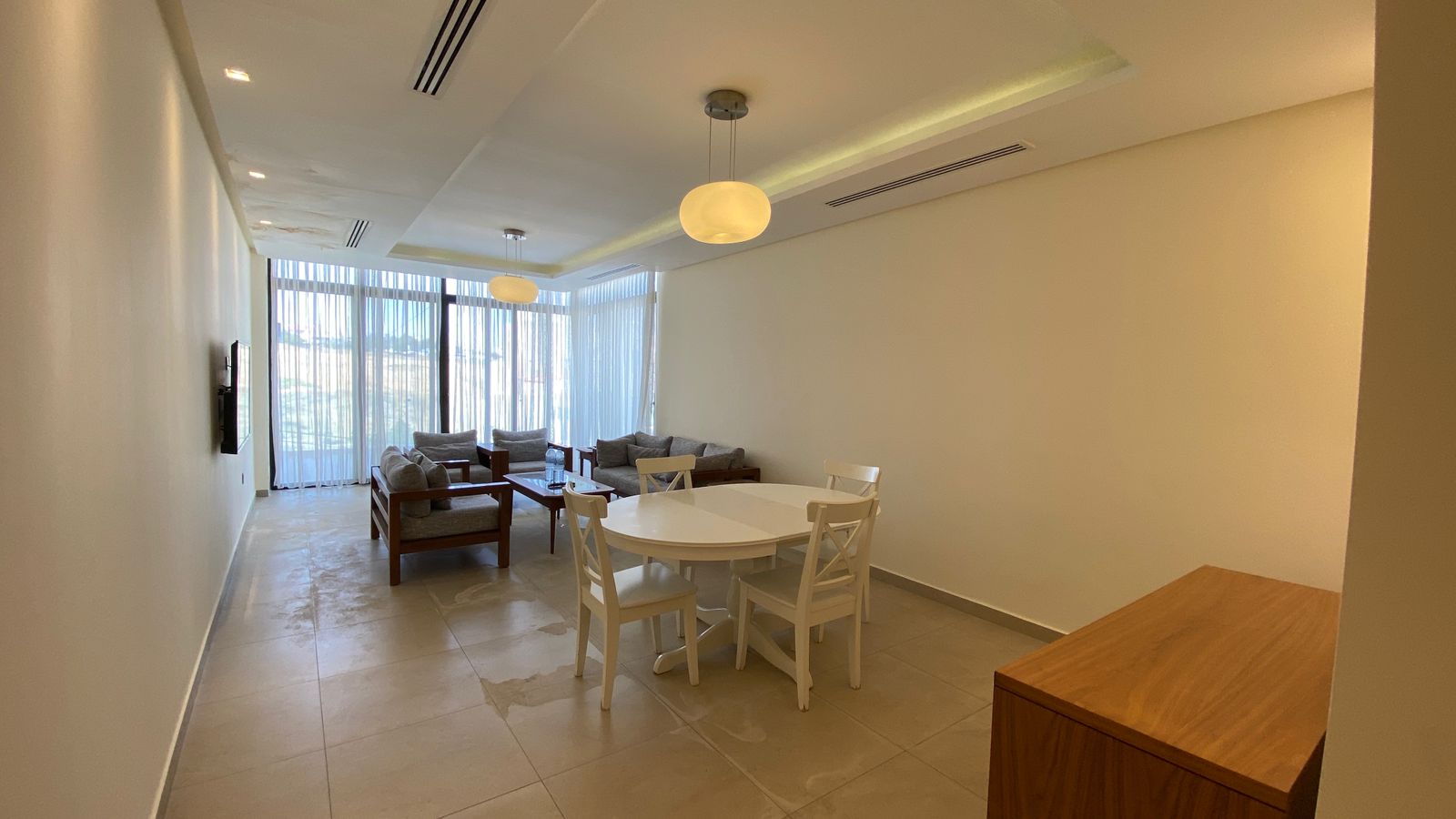 a316d327-dae5-4cb9-8b50-02706a445698 Bright living and dining space: white round table with four white chairs, gray-cushioned wooden sofa set, and large windows with sheer curtains.