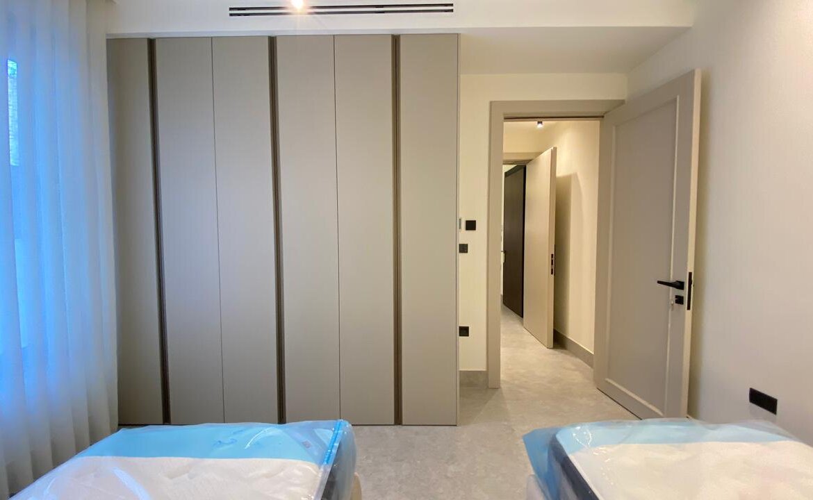 Modern bedroom with tall built-in wardrobes along the wall and two neatly wrapped beds in the foreground; a hallway leads to a door at right.