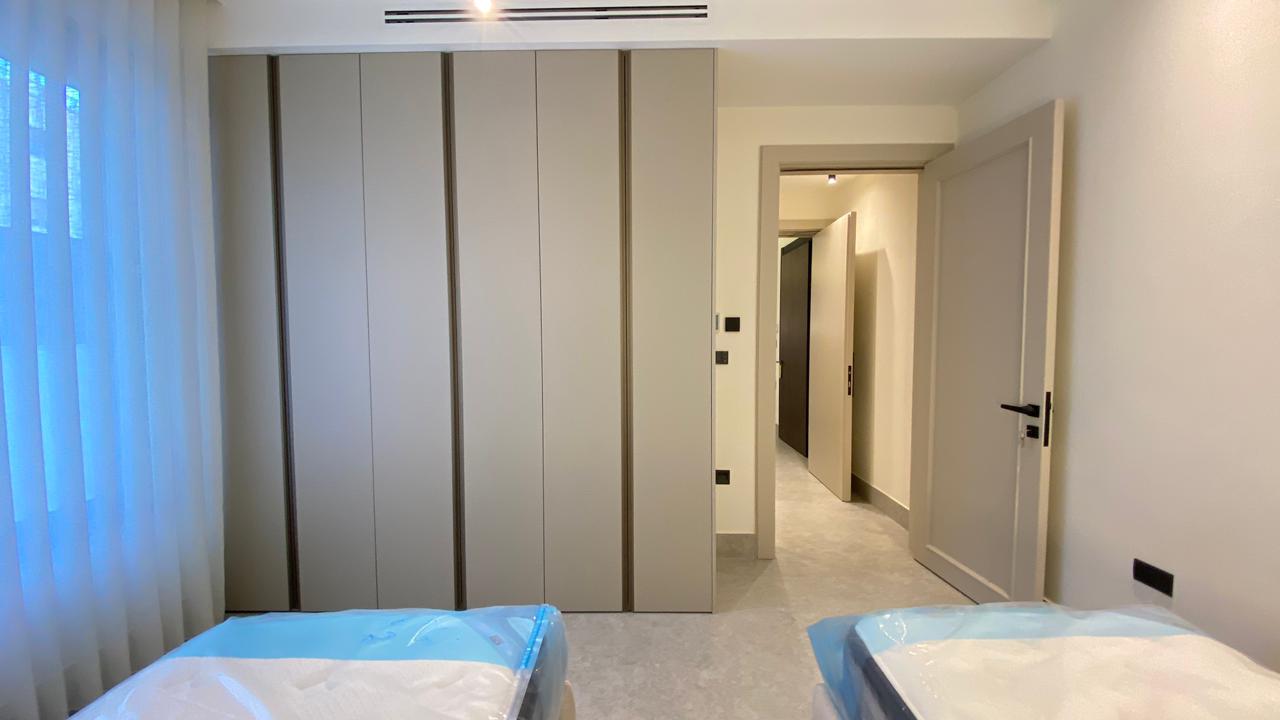 Modern bedroom with tall built-in wardrobes along the wall and two neatly wrapped beds in the foreground; a hallway leads to a door at right.