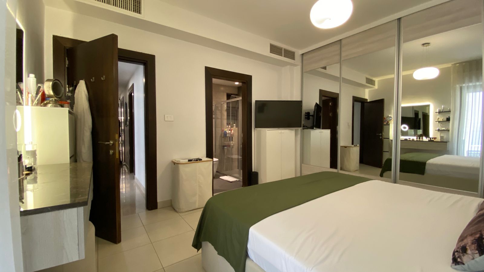 a473e00e-f21a-4cfa-8ce0-3a88036c7cd1 Hotel bedroom with a large bed in white linens and a green bed runner, mirrored wardrobe doors, and a wall-mounted TV.